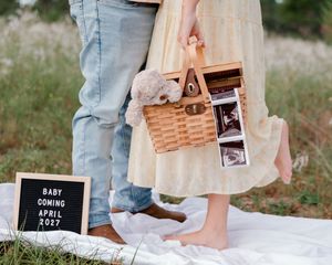 Couple holding a wicker basket with ultrasound photos and a letterboard announcing baby coming April 2027.