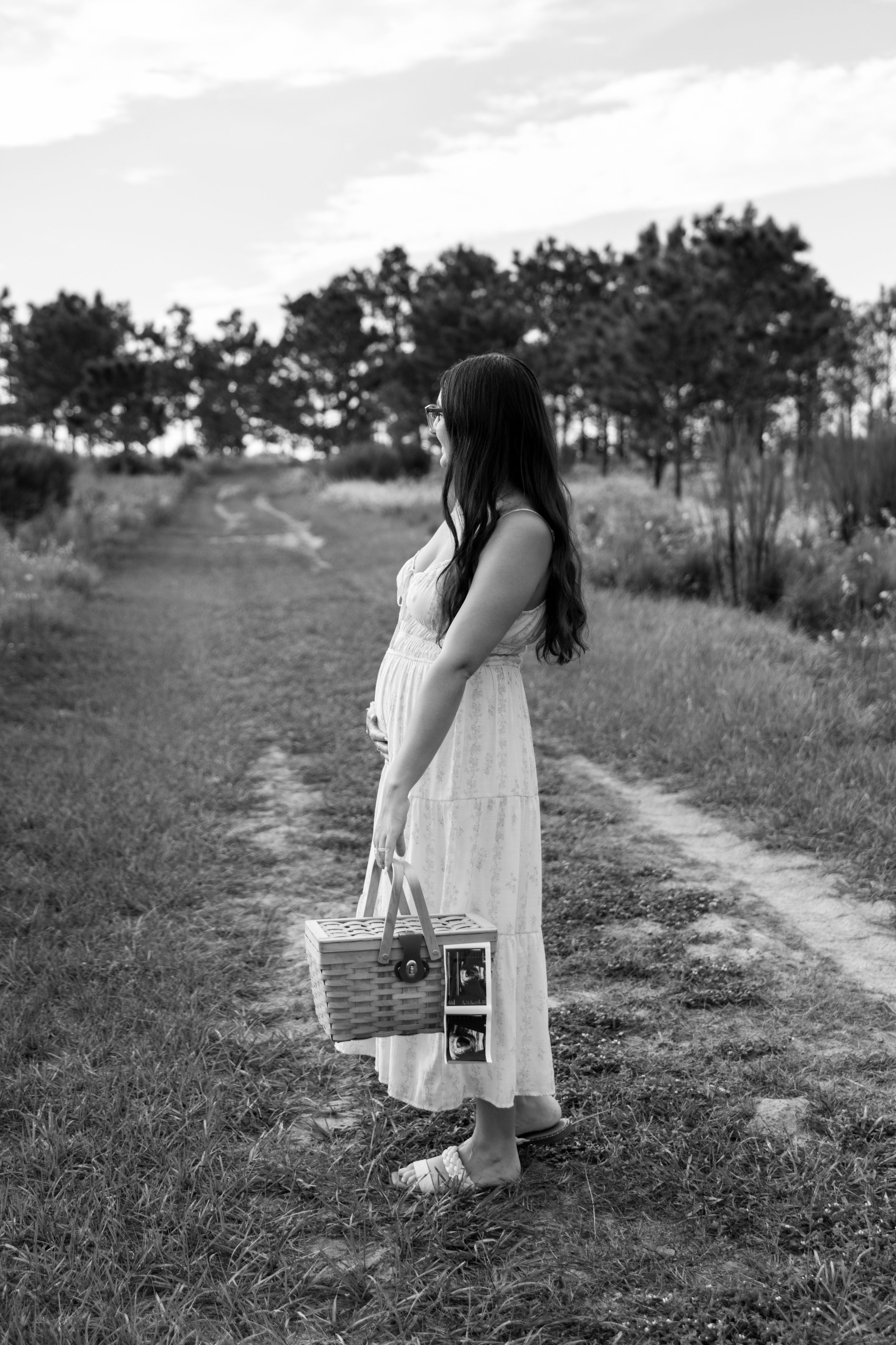 Black and white photo of a pregnant woman in a white dress holding a picnic basket on a rural path surrounded by trees.