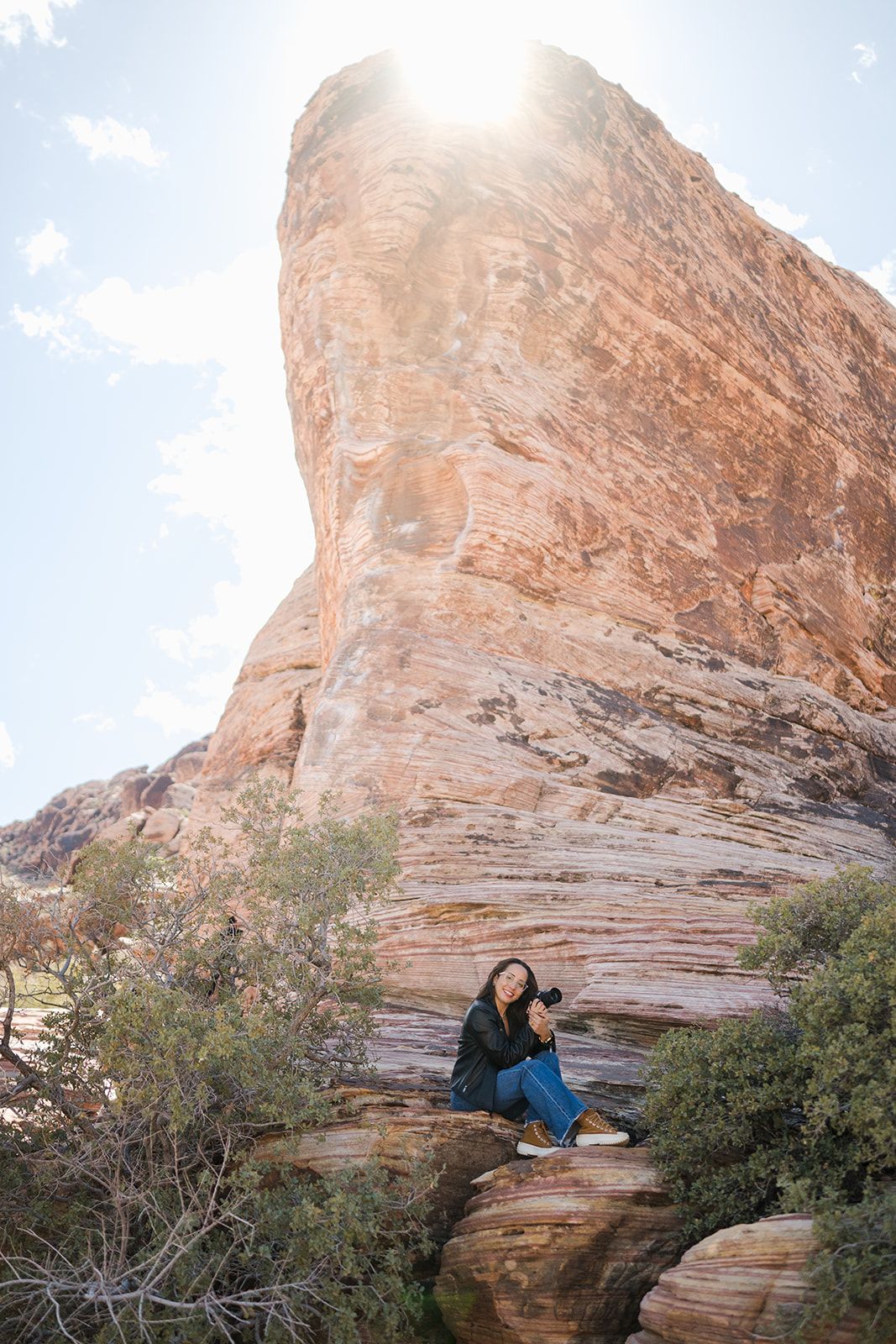 Person sitting on red rock formation with majestic cliff and sunburst in background.