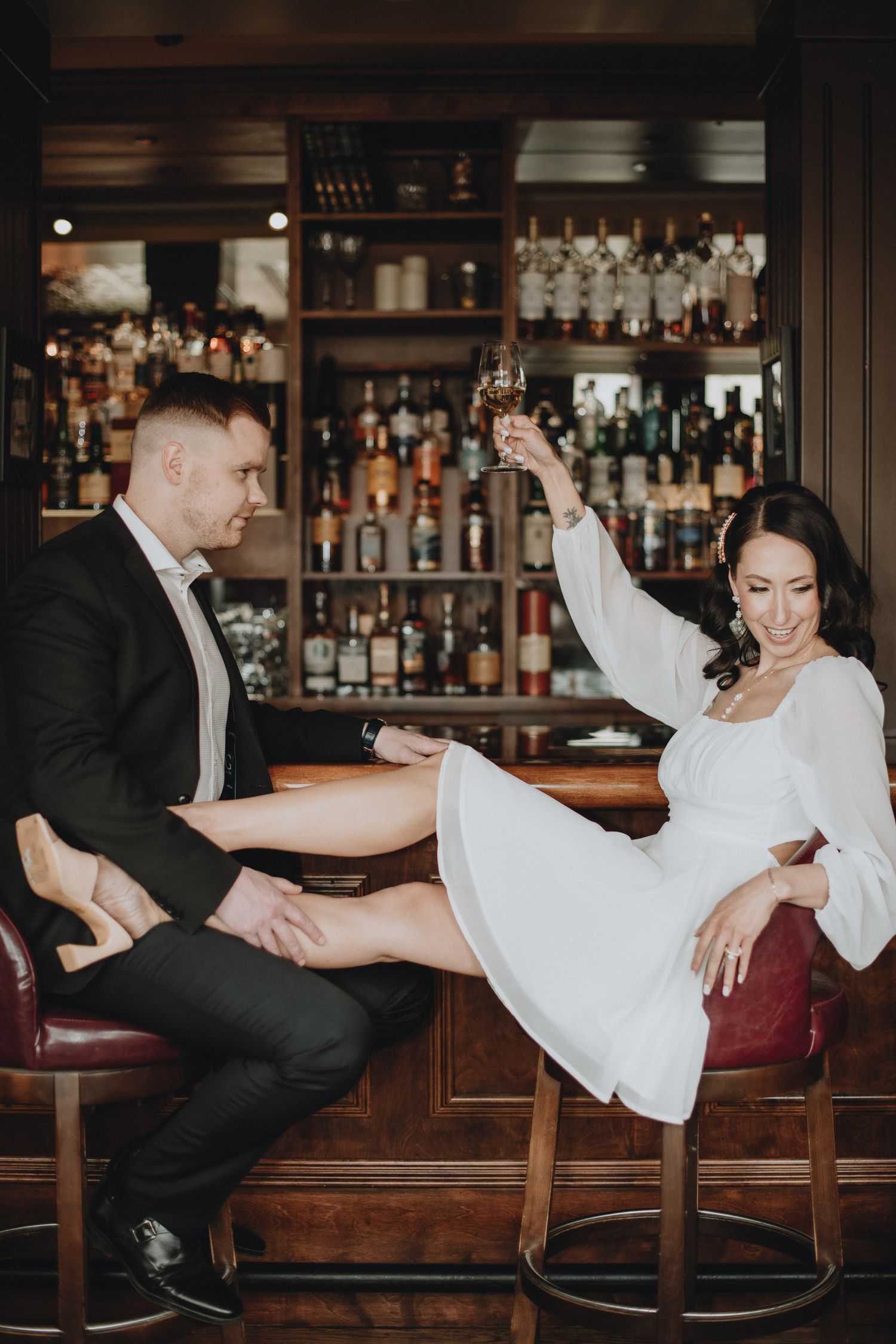 Couple enjoying drinks at an elegant bar with woman in white dress playfully lifting leg while seated on bar stool.