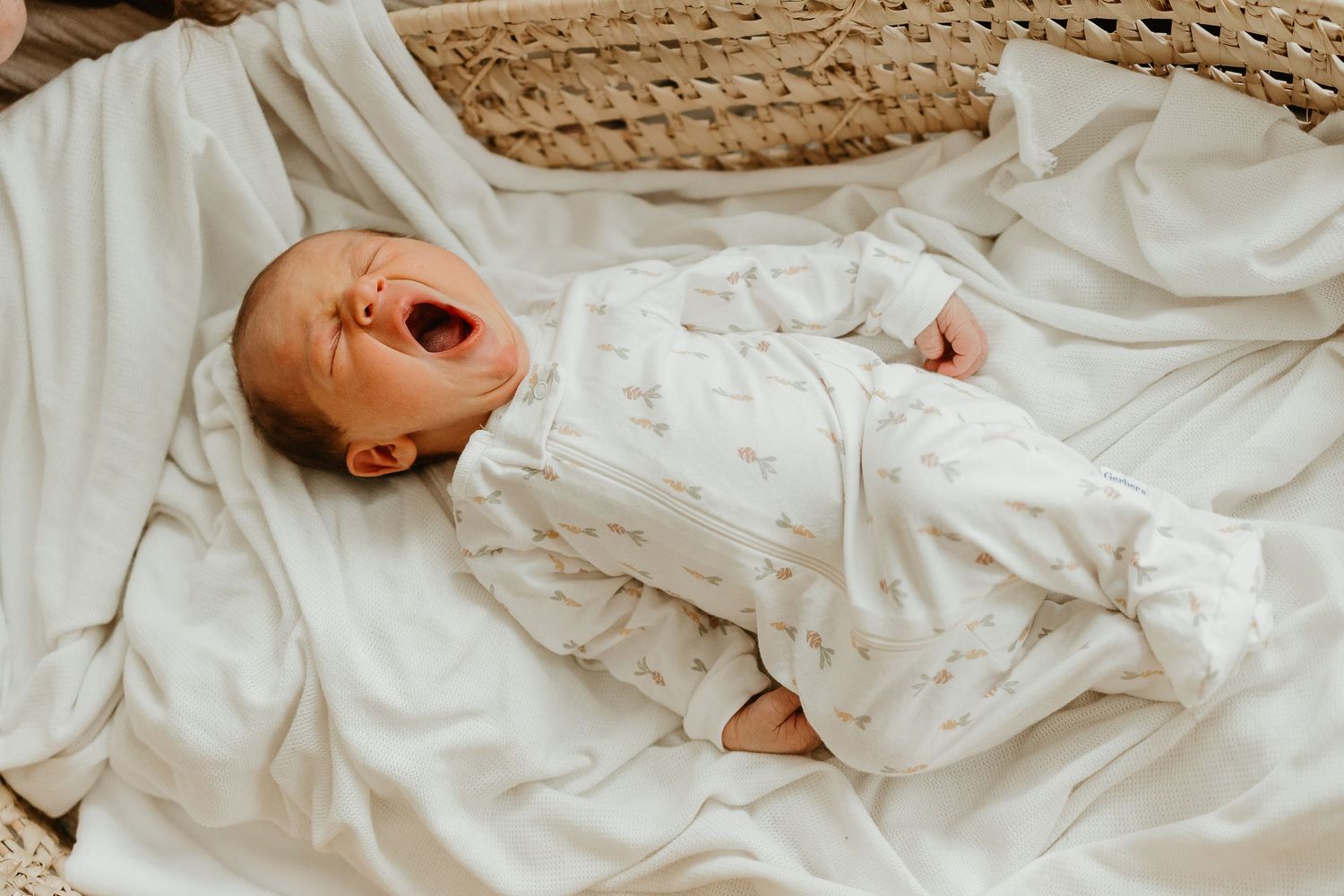 A yawning baby wearing a white floral outfit lies on soft white bedding.