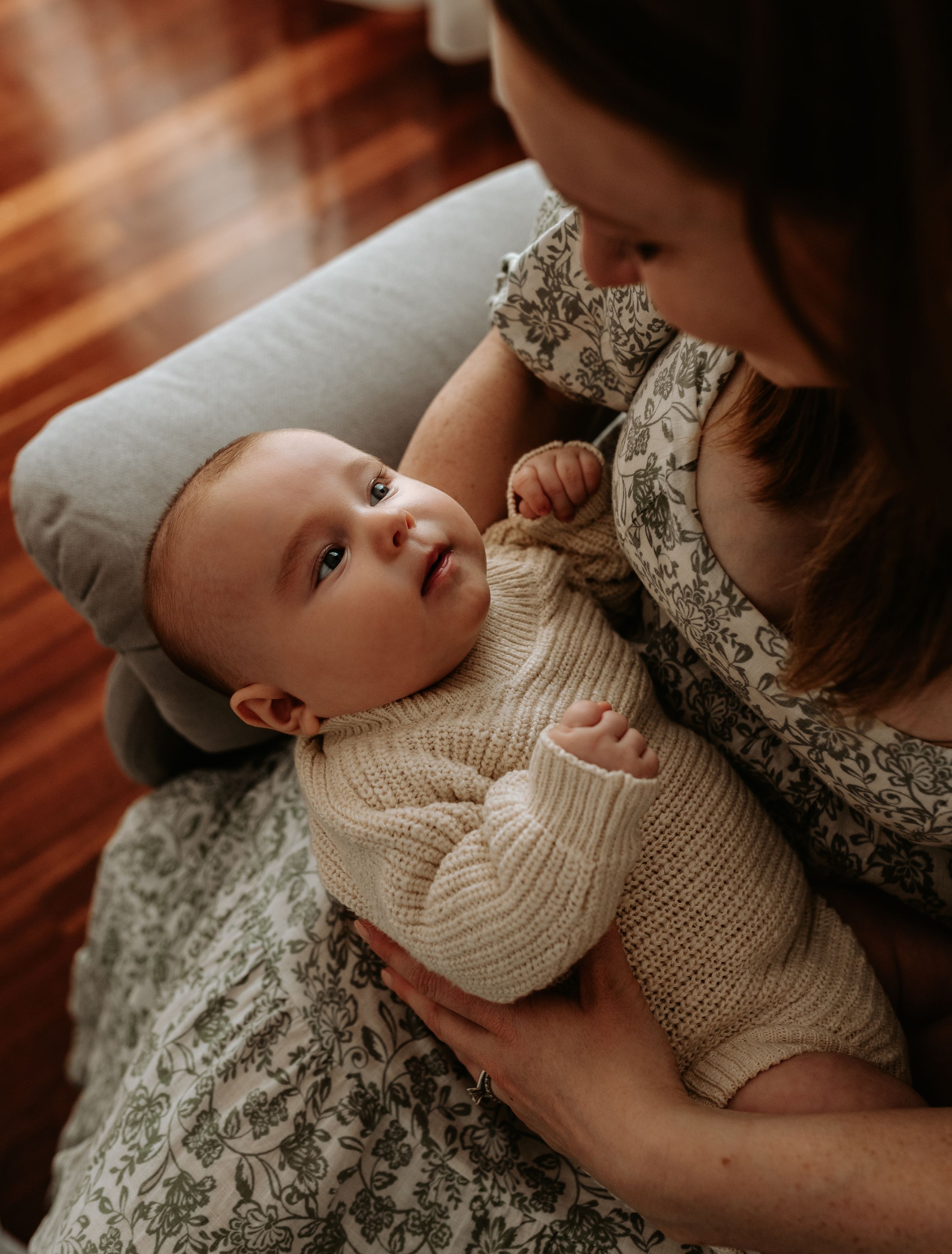 A baby wearing a cream knit sweater lies on hardwood floor while being comforted by someone in a floral dress.