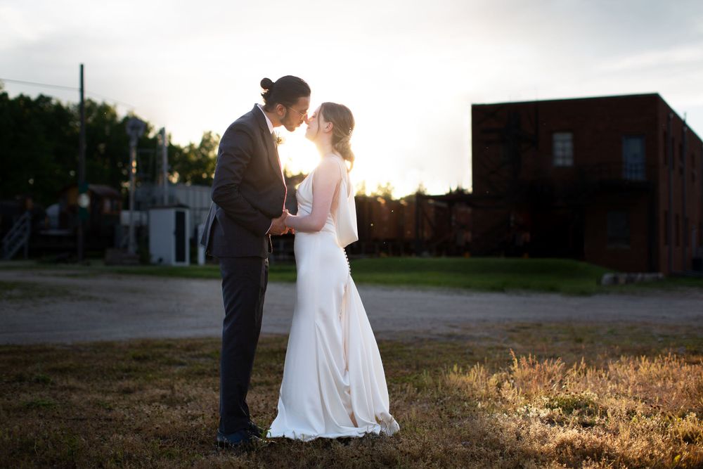 A wedding couple shares an intimate moment at sunset near a rustic building.