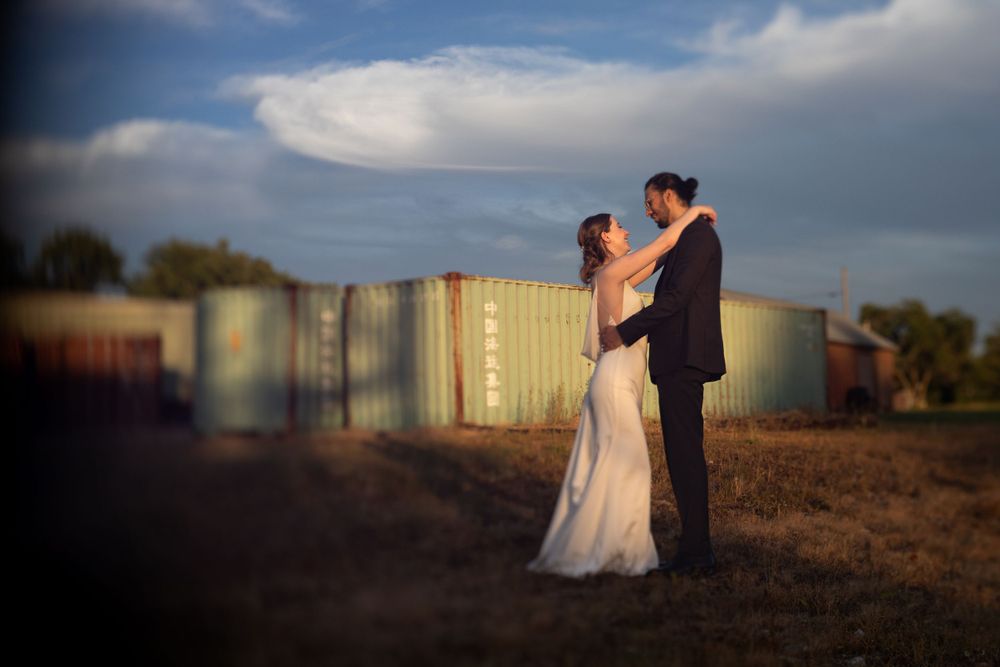 A couple embraces in an outdoor setting at sunset with industrial storage tanks in the background.