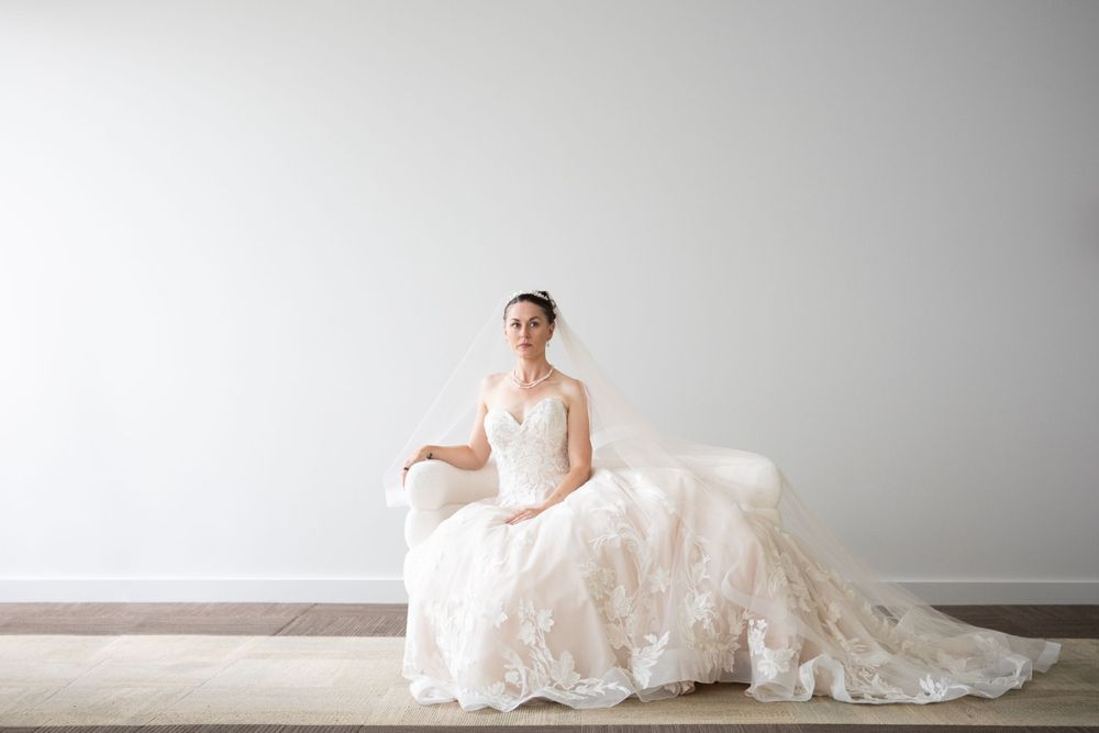 A bride in an elegant white wedding gown with a flowing train poses in a minimalist studio setting.