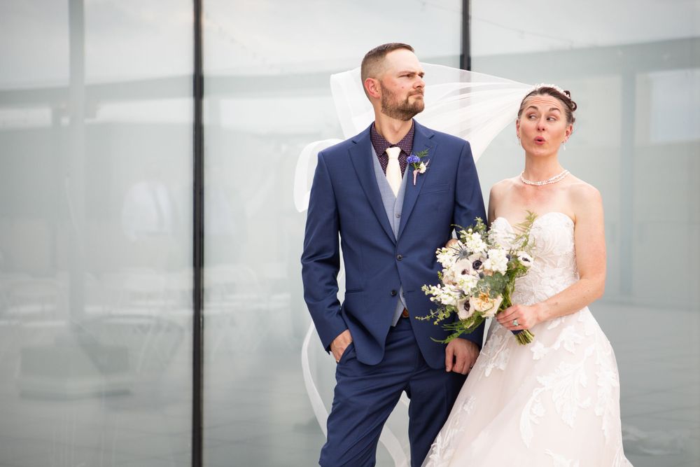 A couple poses together against a modern grey backdrop holding a white floral bouquet.