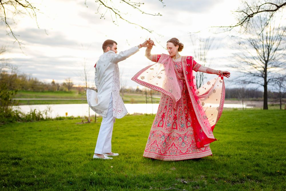 A couple in traditional Indian attire dances joyfully in a green field by a lake.