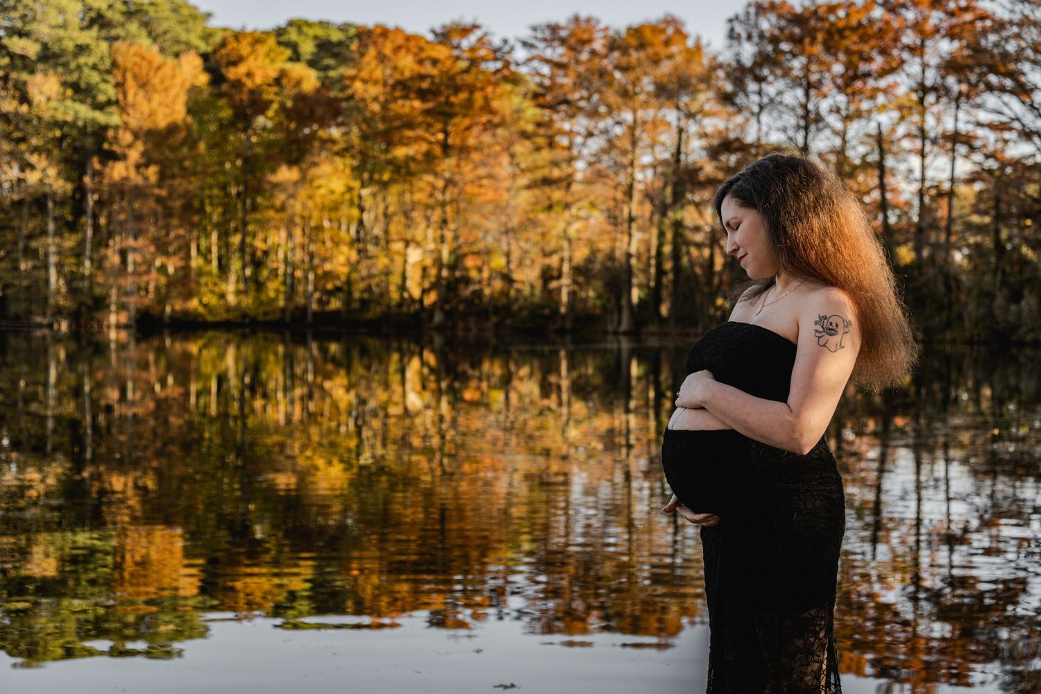 Intimate maternity photo of couple embracing by a serene lake surrounded by pine trees.