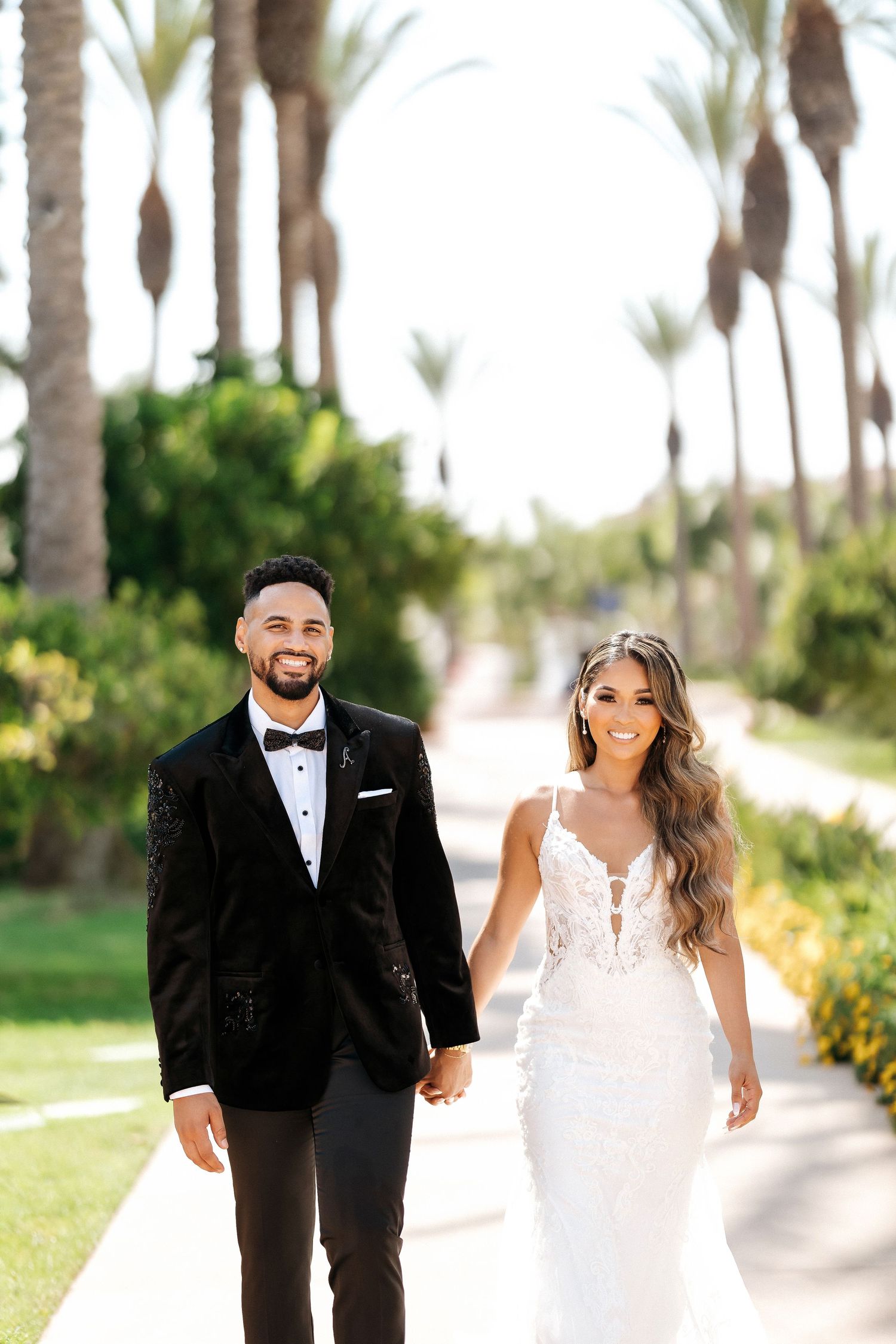 Wedding couple walks palm-lined path in elegant attire during outdoor photo session.