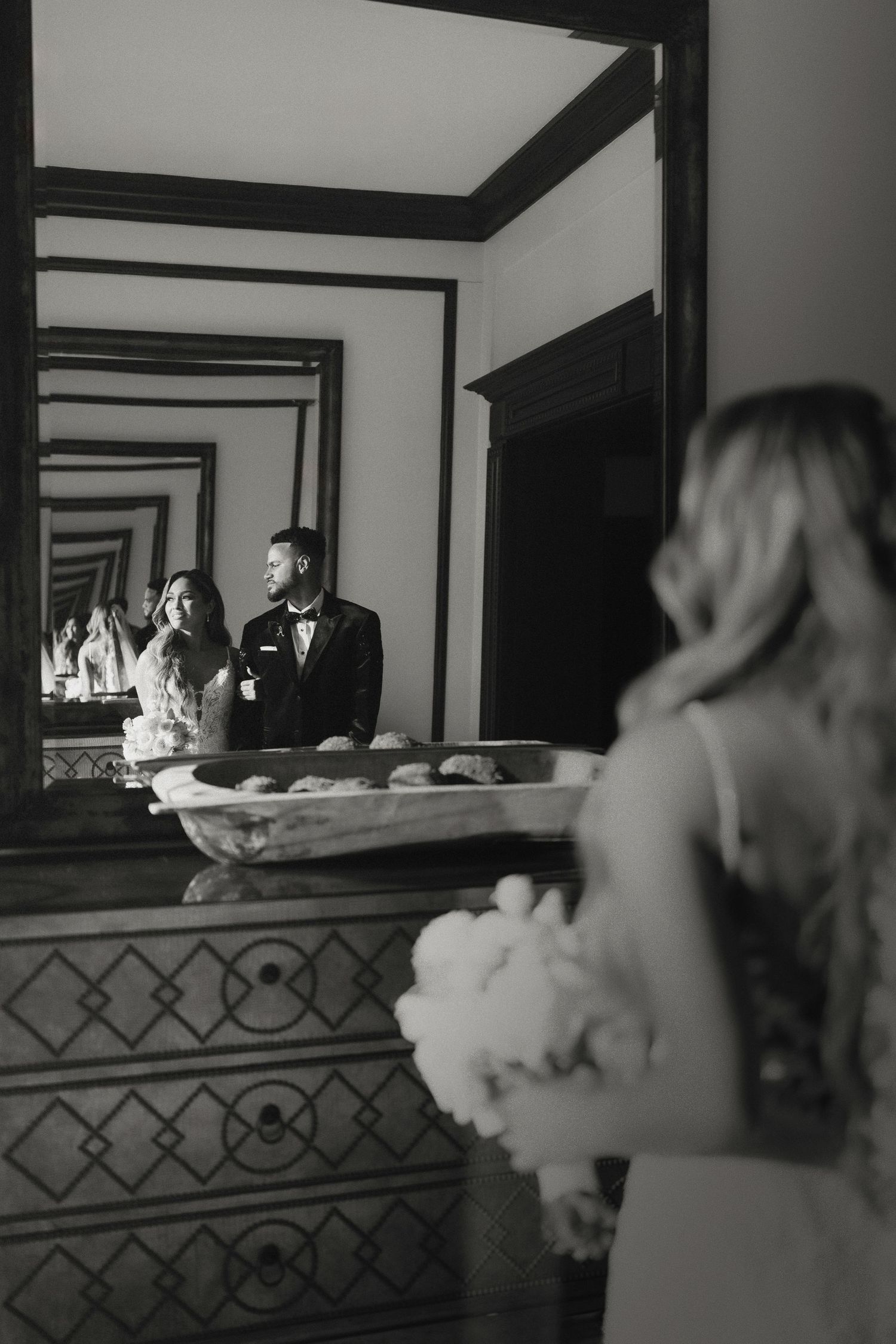 Black and white wedding photo showing couple reflected in ornate mirror during preparation.