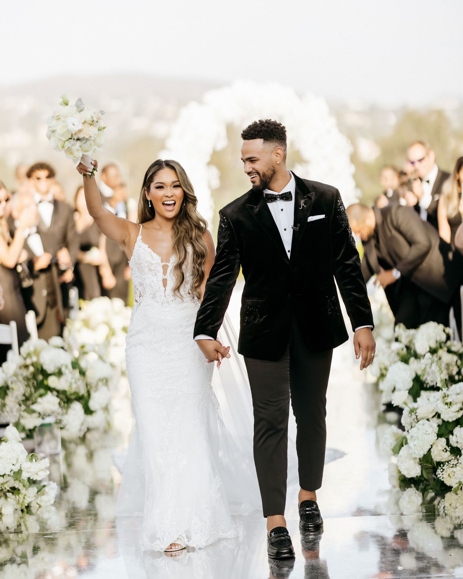 Wedding couple walks down aisle surrounded by white flowers and celebrating guests.