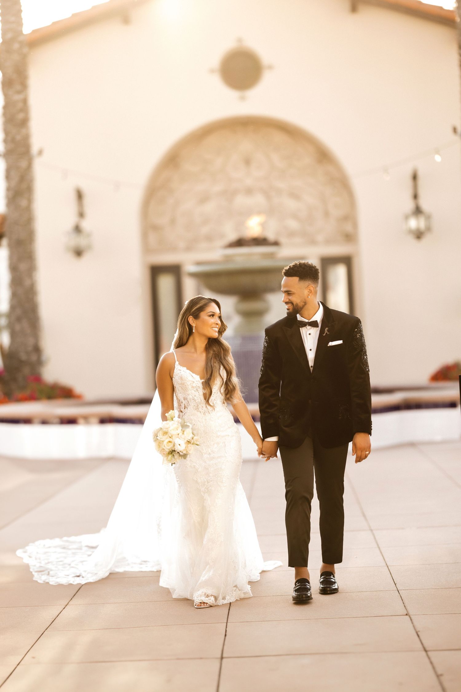 Newlyweds walk hand in hand outside Mediterranean-style venue with arched doorway.
