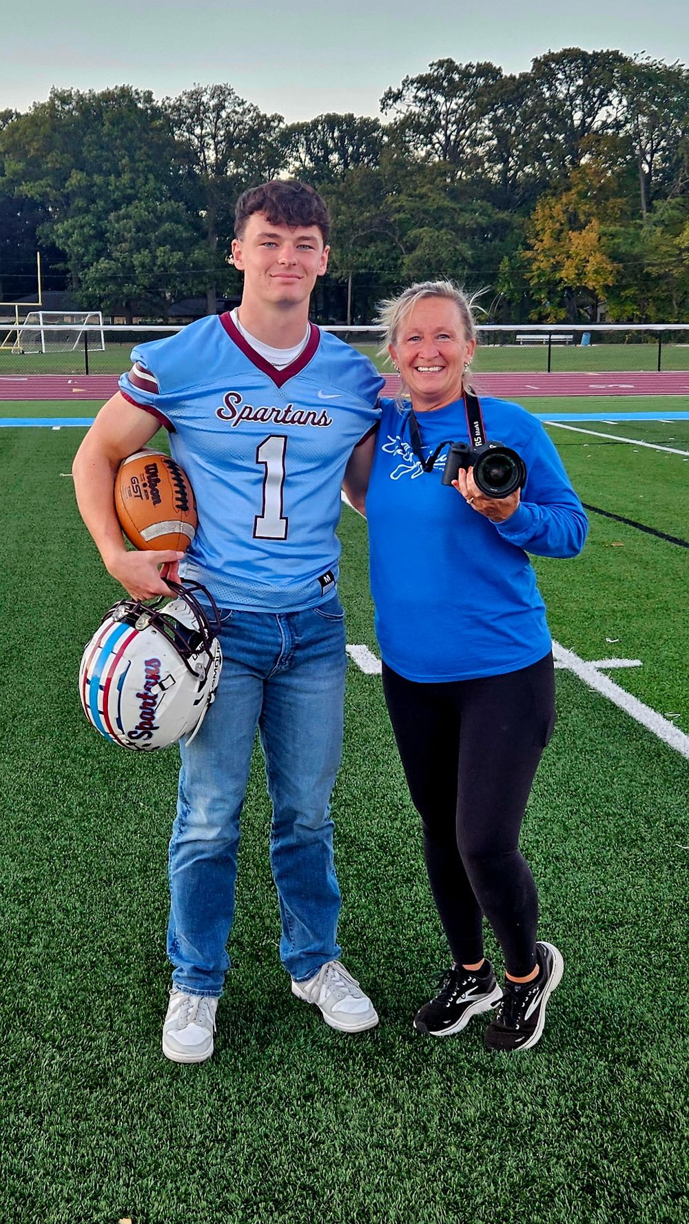 Photographer and her senior client pose together on a football field holding football equipment and a camera.