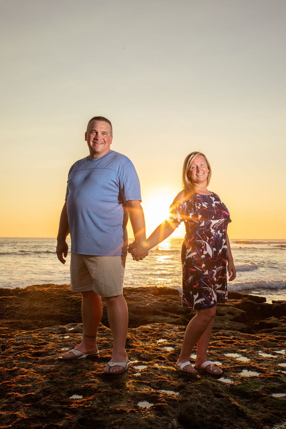 Two people pose playfully on a beach at sunset with ocean waves and rocky shore in the background.
