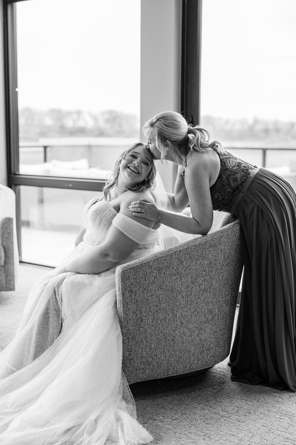 A black and white wedding photo of the bride and her mother sharing a special moment by large windows.
