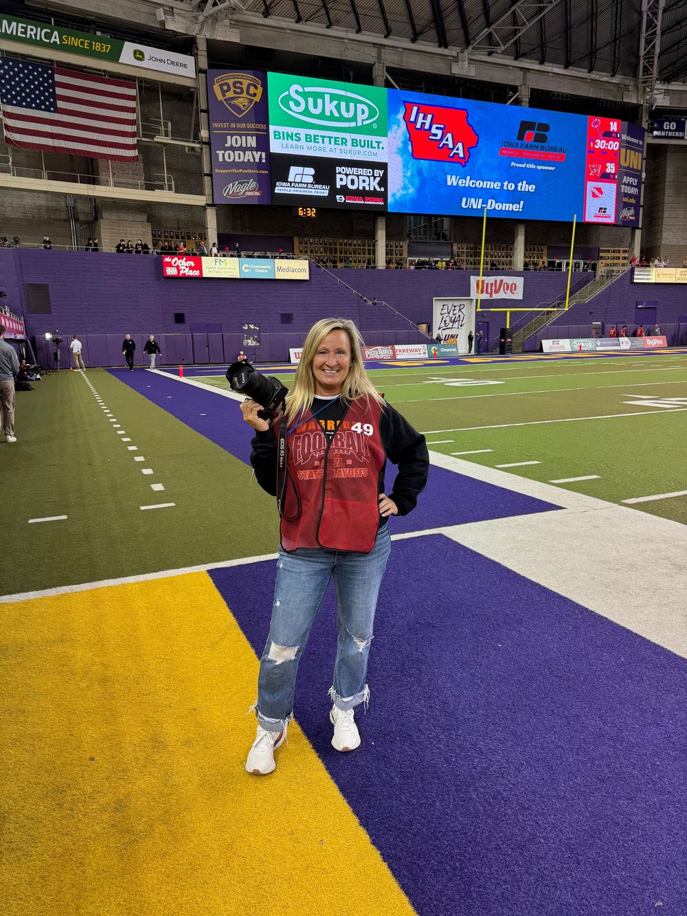 Photographer stands on a football field with purple and yellow end zone markings inside an indoor stadium with American flag visible.