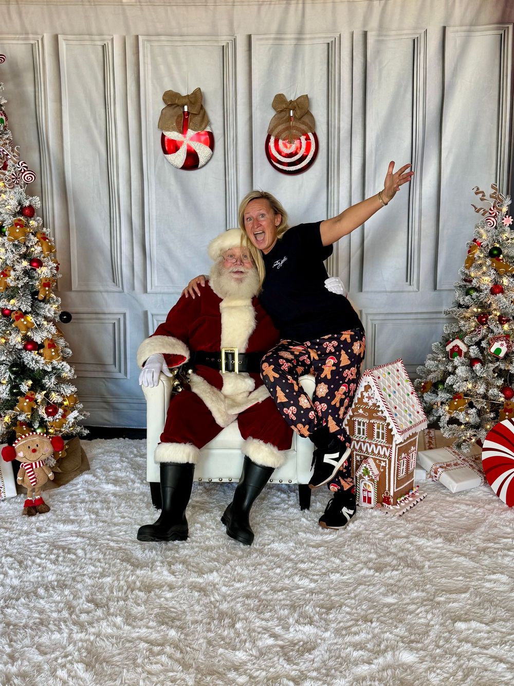 Truly Juli poses with Santa at a Christmas scene with decorated trees, a gingerbread house, and holiday wall decor.