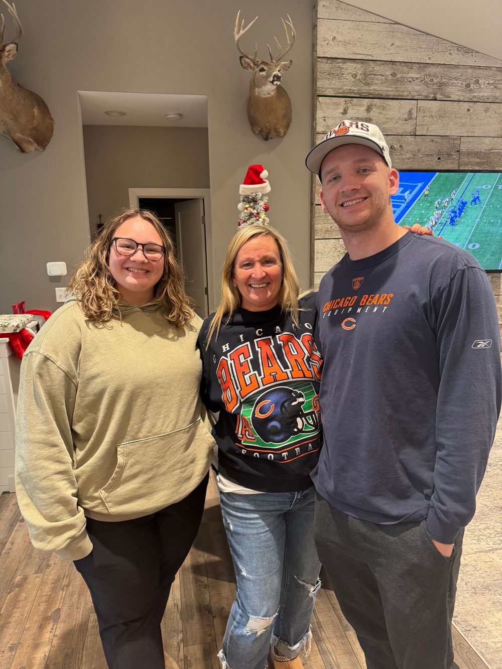 Mother and her adult children pose in front of mounted deer heads and a rustic wood wall with a football game on TV.