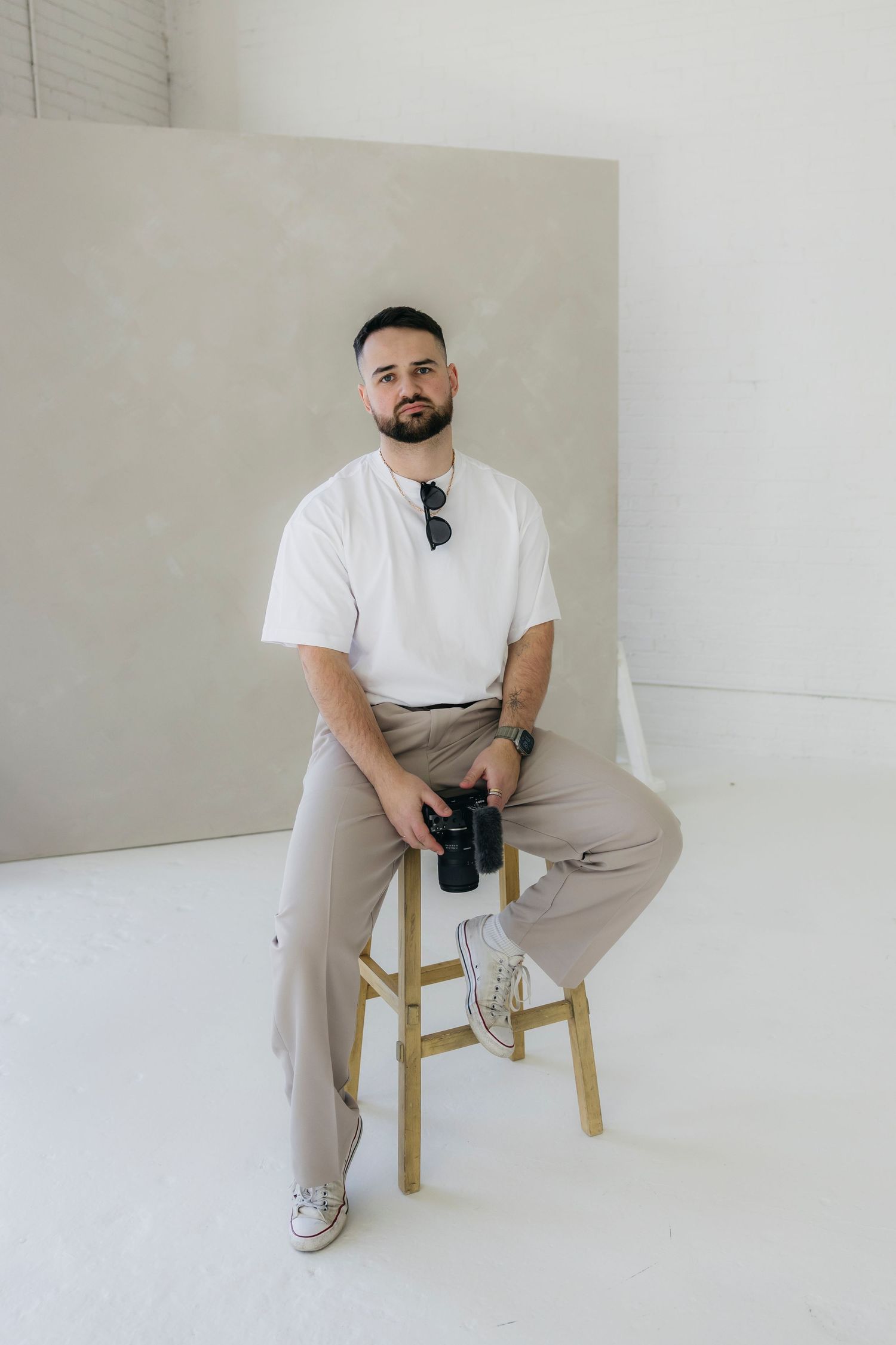 Person in casual white t-shirt and khaki pants sitting on wooden stool against white background.