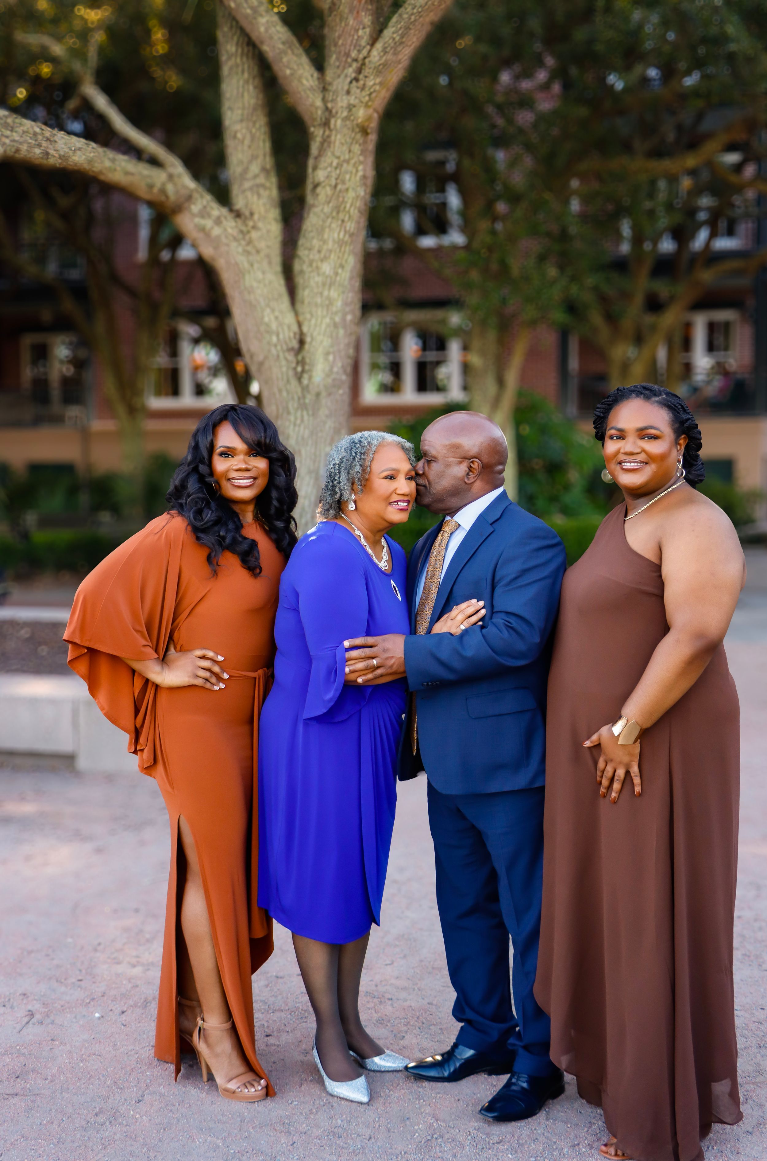 Family of four smiling outdoors in Downtown Charleston, South Carolina, parents sharing a tender moment while daughters stand beside them.