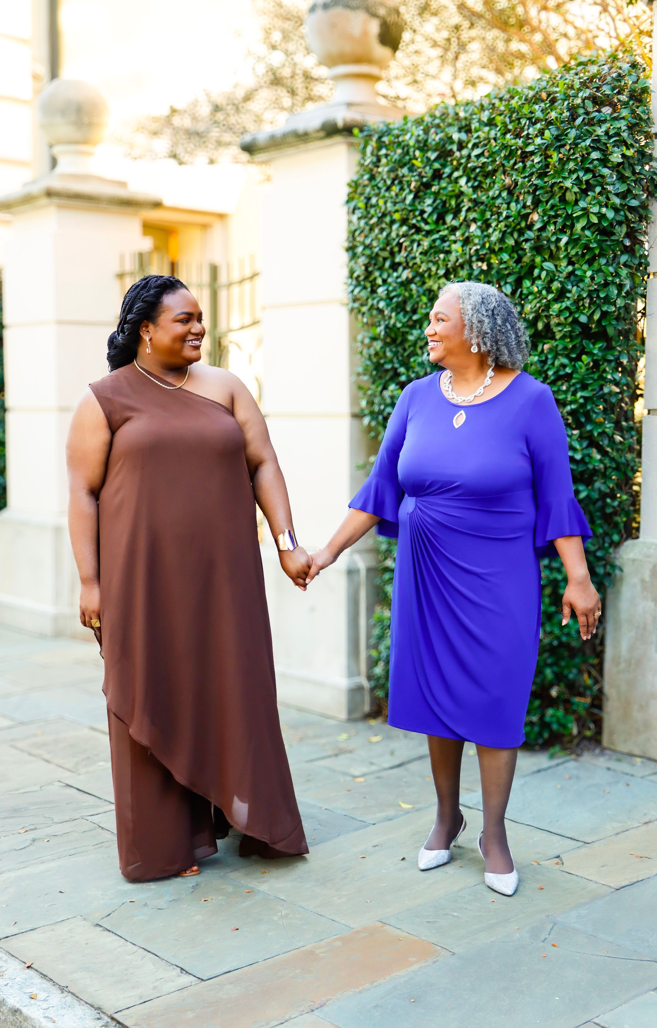 mother and daughter holding hands and smiling at each other, wearing brown asymmetric gown and blue dress outdoors.