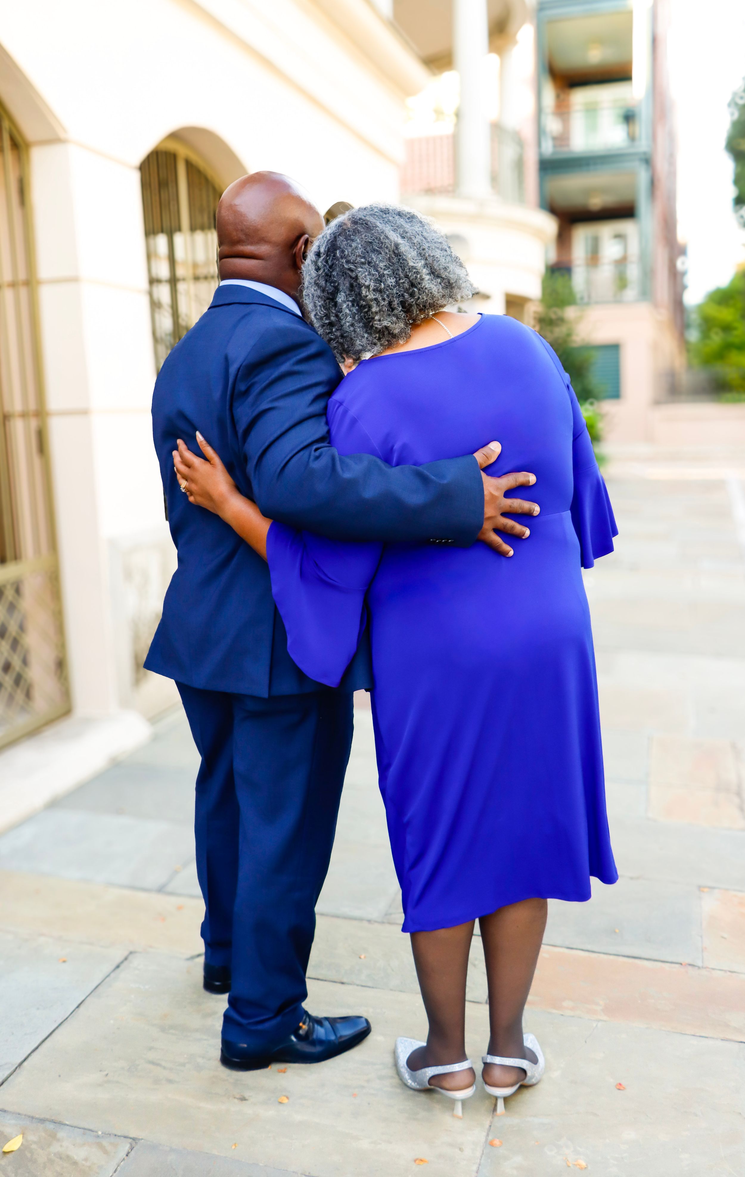 Couple embracing from behind outdoors, man in navy suit and woman in royal blue dress with gray hair.
