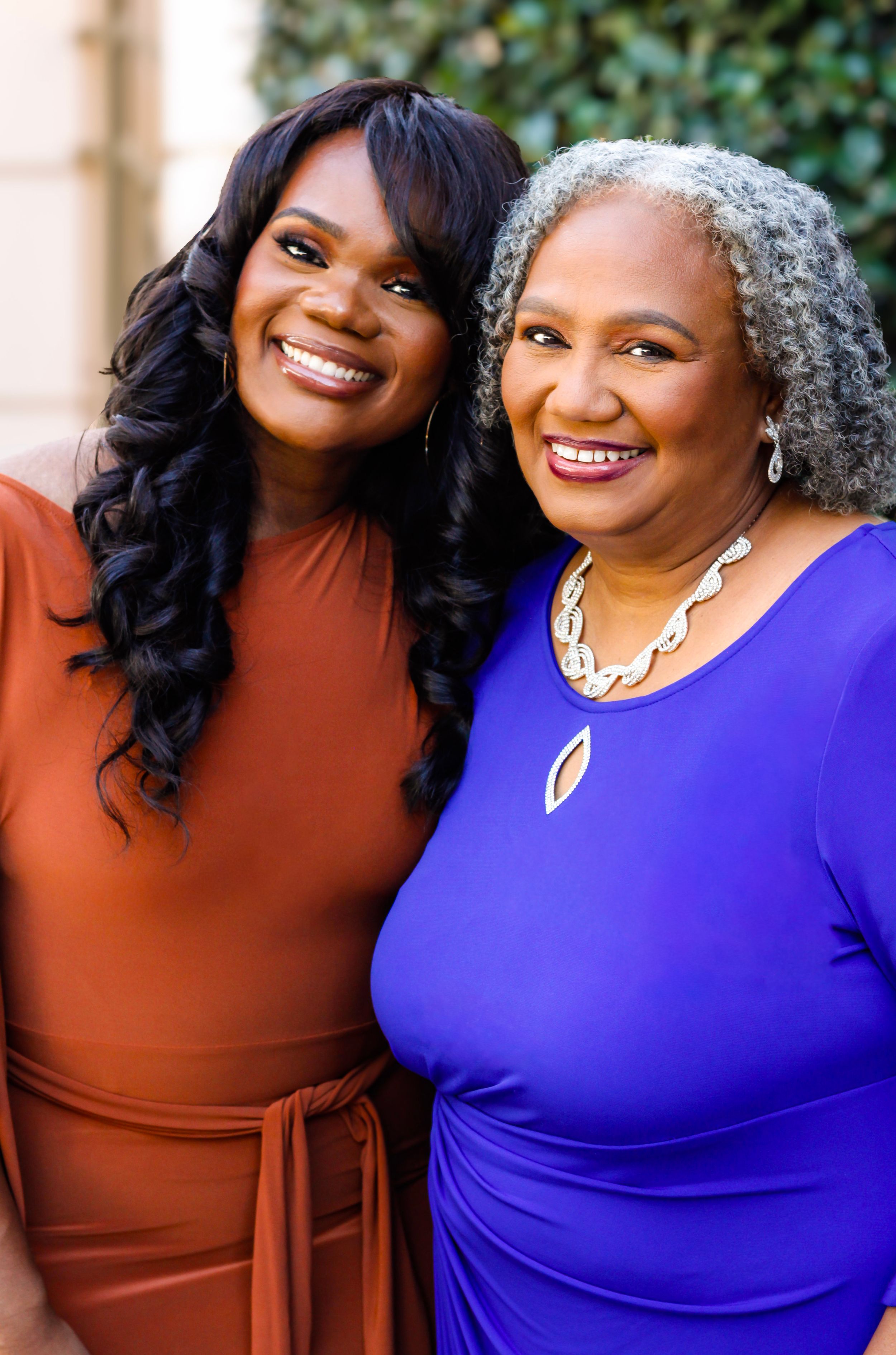 Two smiling women posing together, one in rust dress with curly hair, other in purple dress with gray curls.