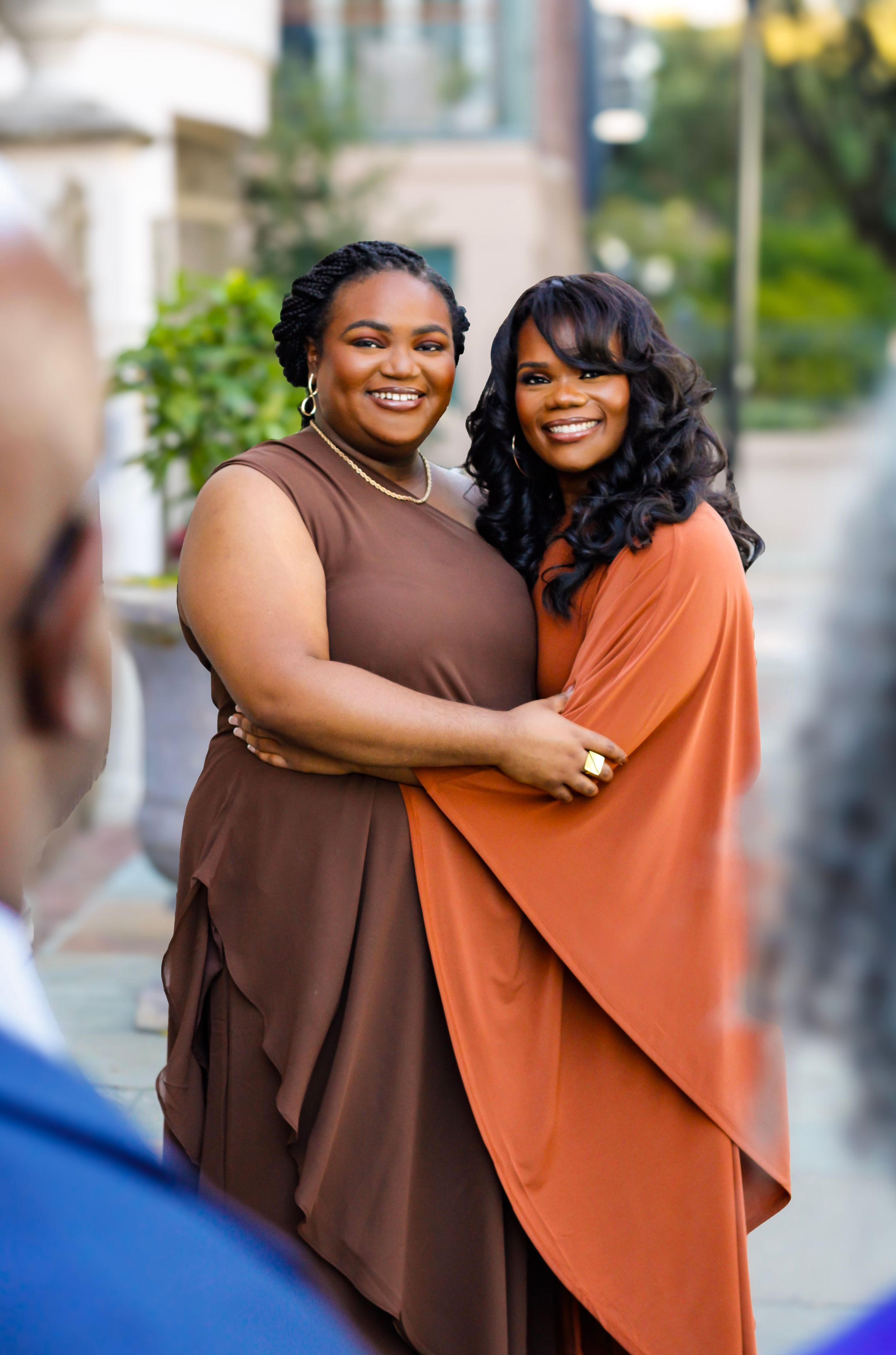 two sisters embracing and smiling outdoors in Downtown Charleston, wearing brown and rust-colored dresses at a formal event.