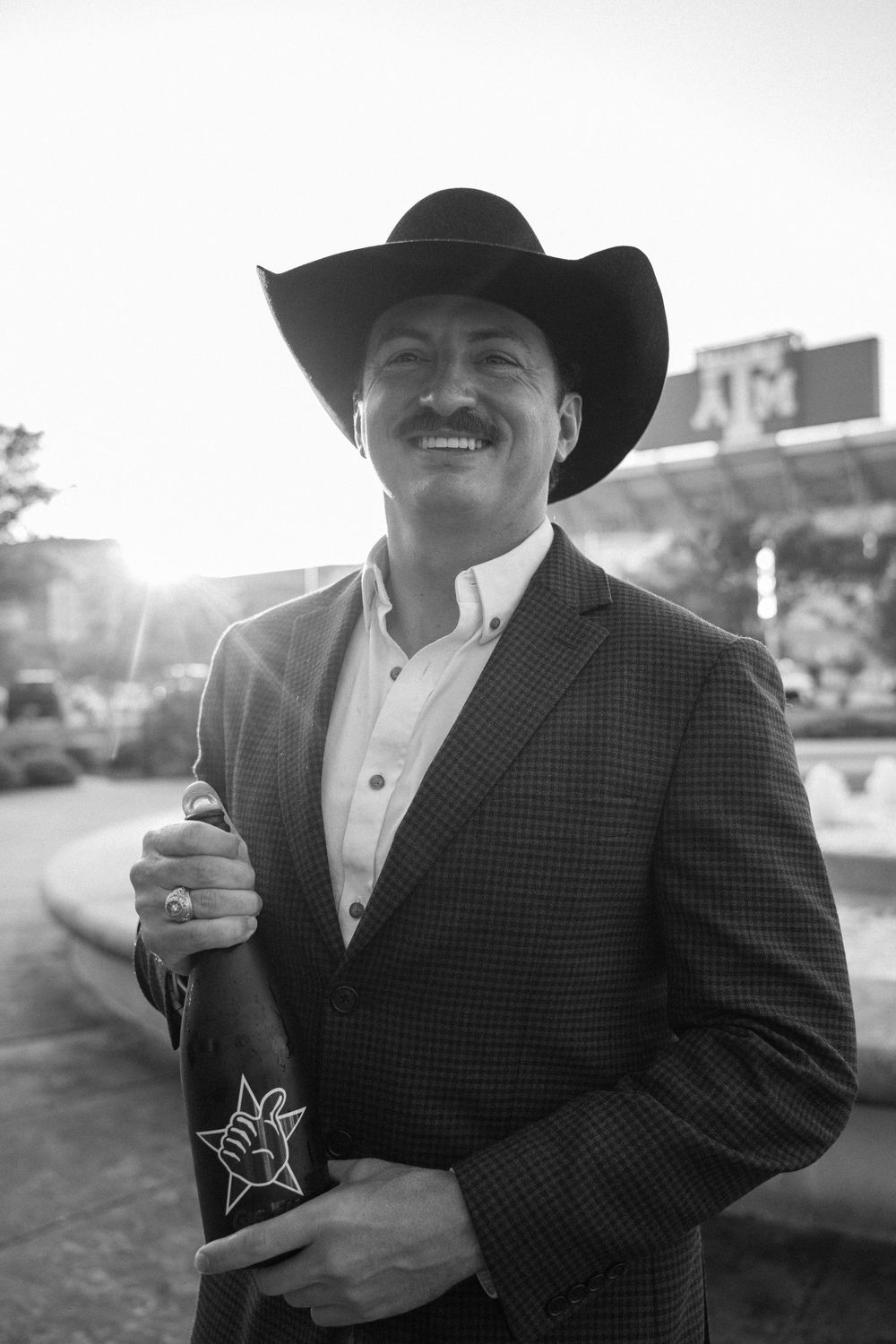 Man in cowboy hat and blazer holding a wine bottle, smiling outdoors in black and white photo.