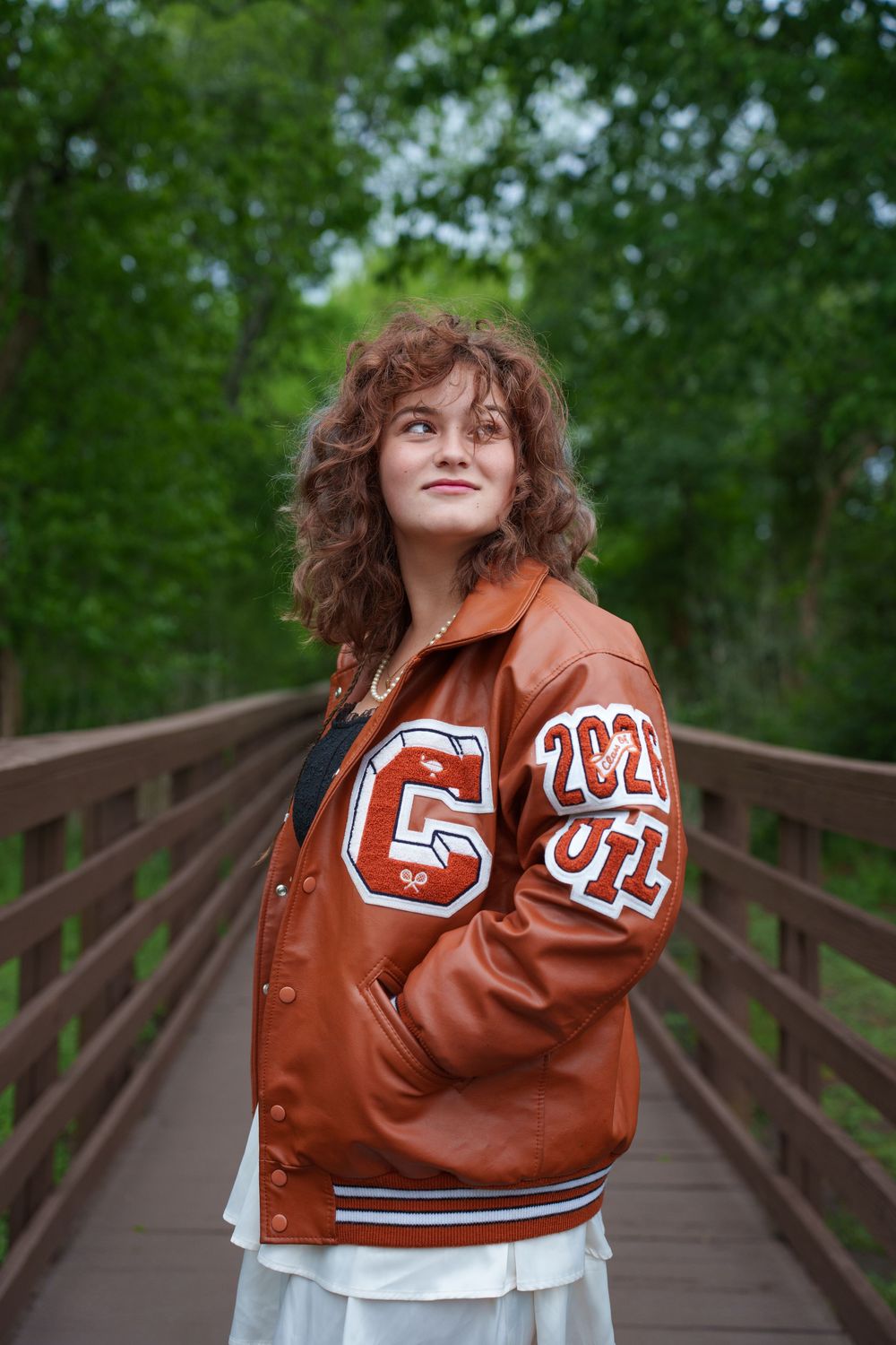 Teen girl in brown varsity jacket with letter C patch stands on wooden bridge surrounded by green trees.