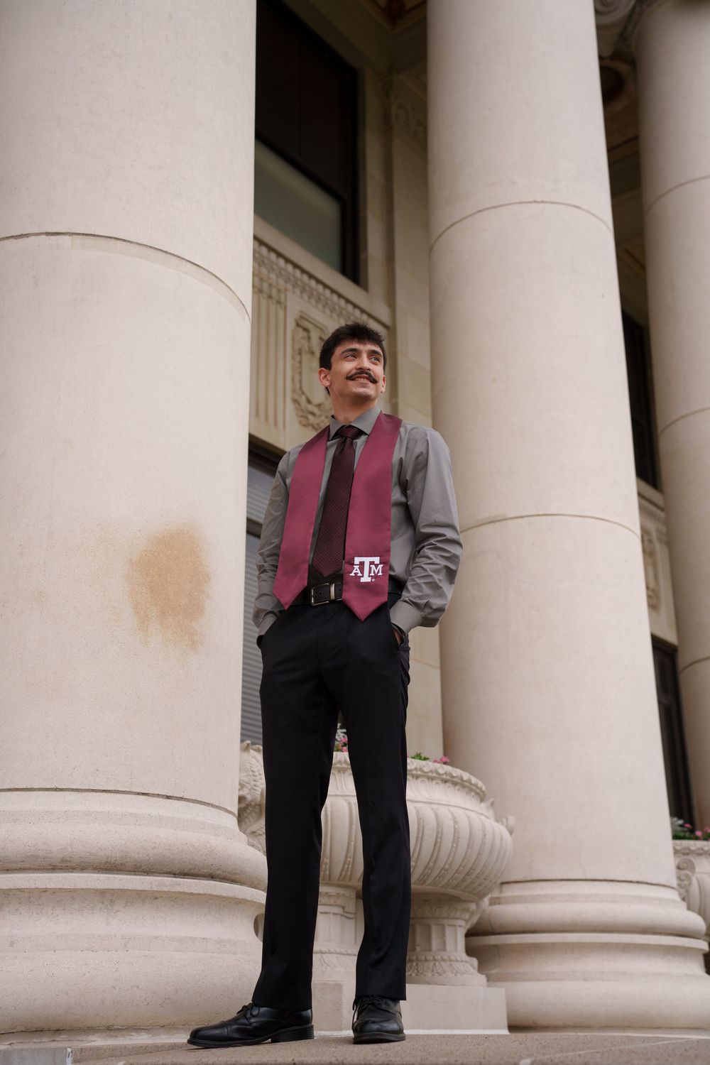 Young man in maroon Texas A&M graduation stole and vest stands between large columns at a university building.