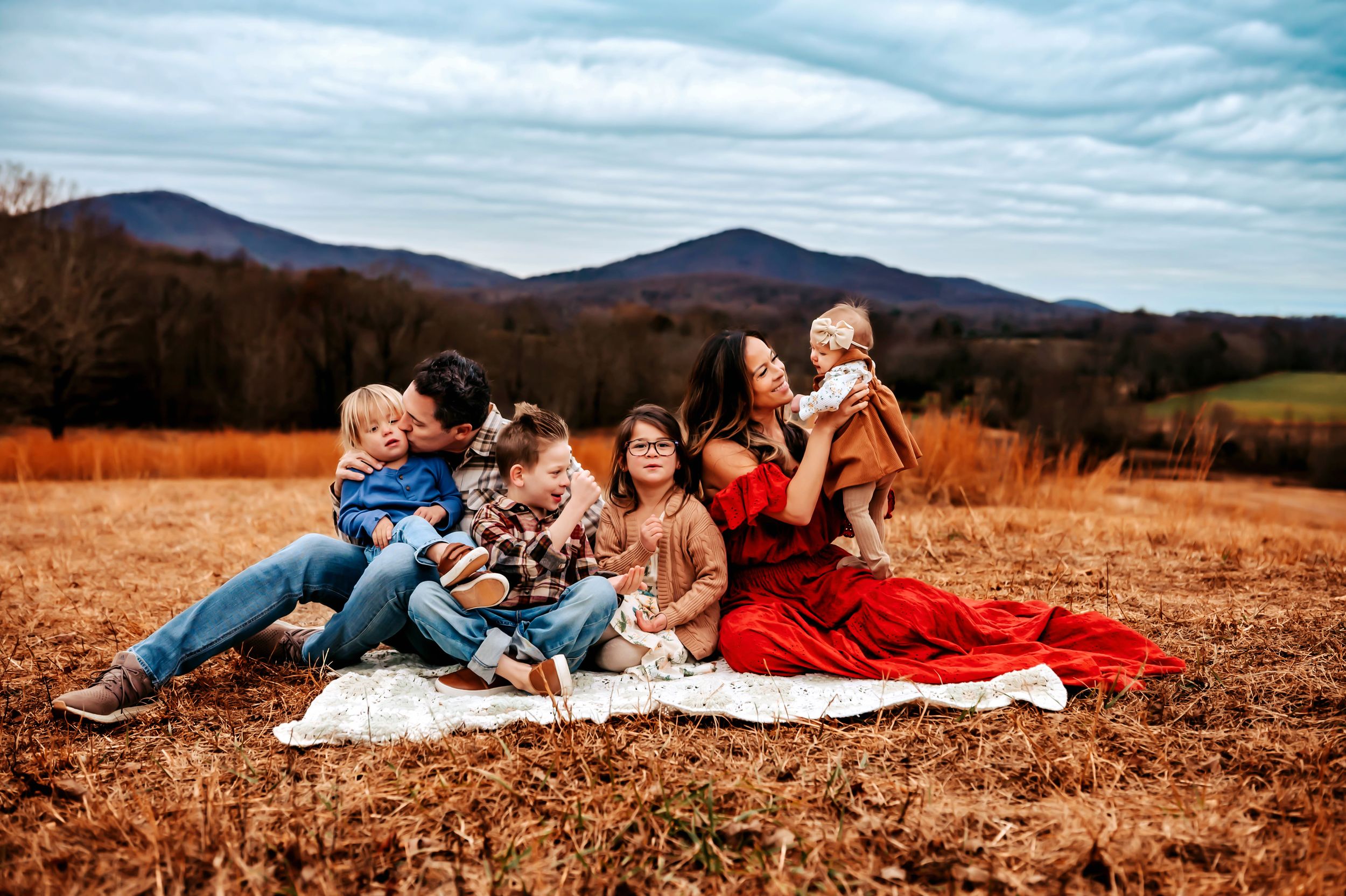 Family members lie together on an autumn blanket in a rustic field with mountains visible in the background.