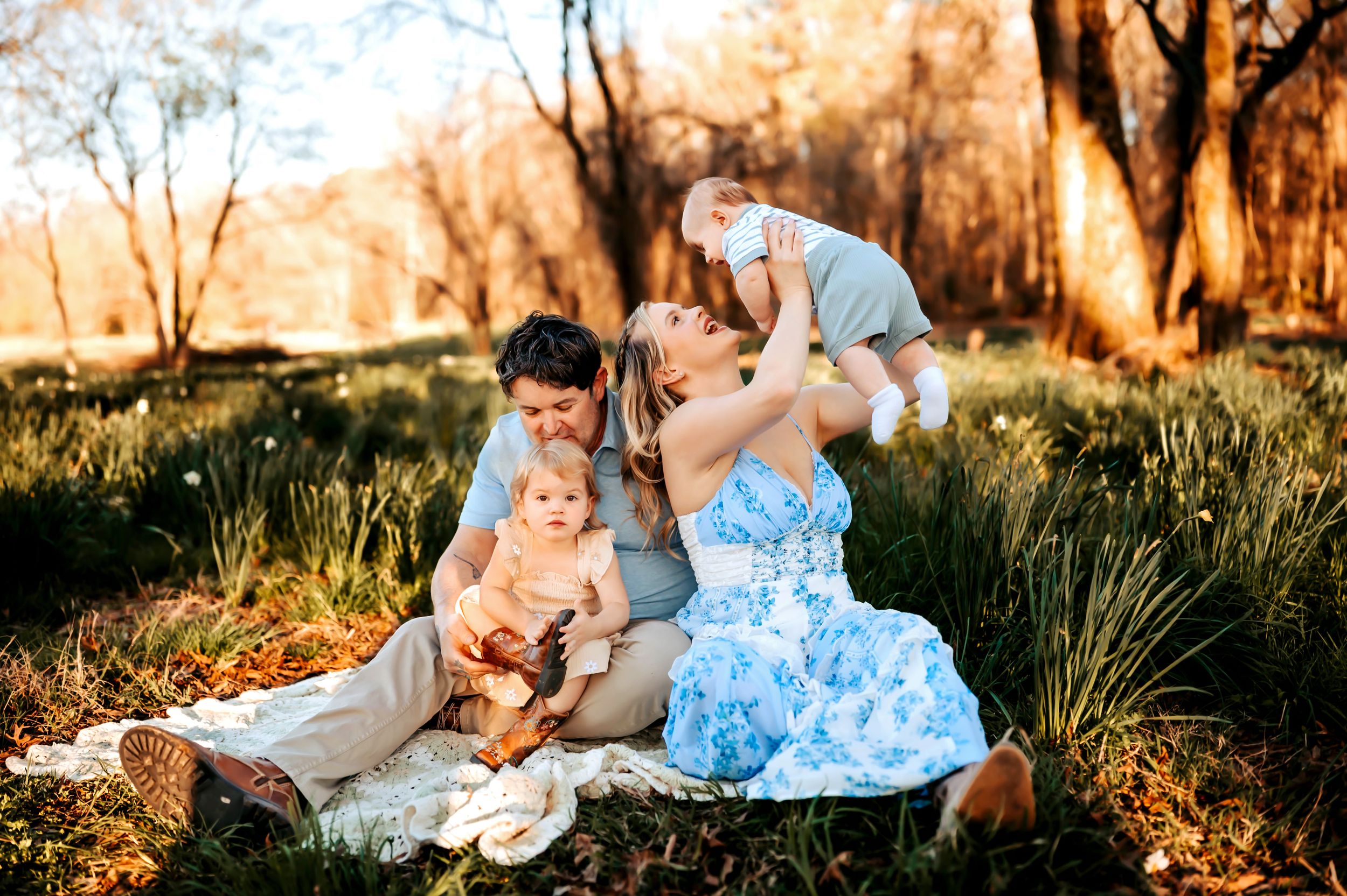 A family sits together on a blanket in a sunlit meadow during golden hour with trees in the background.