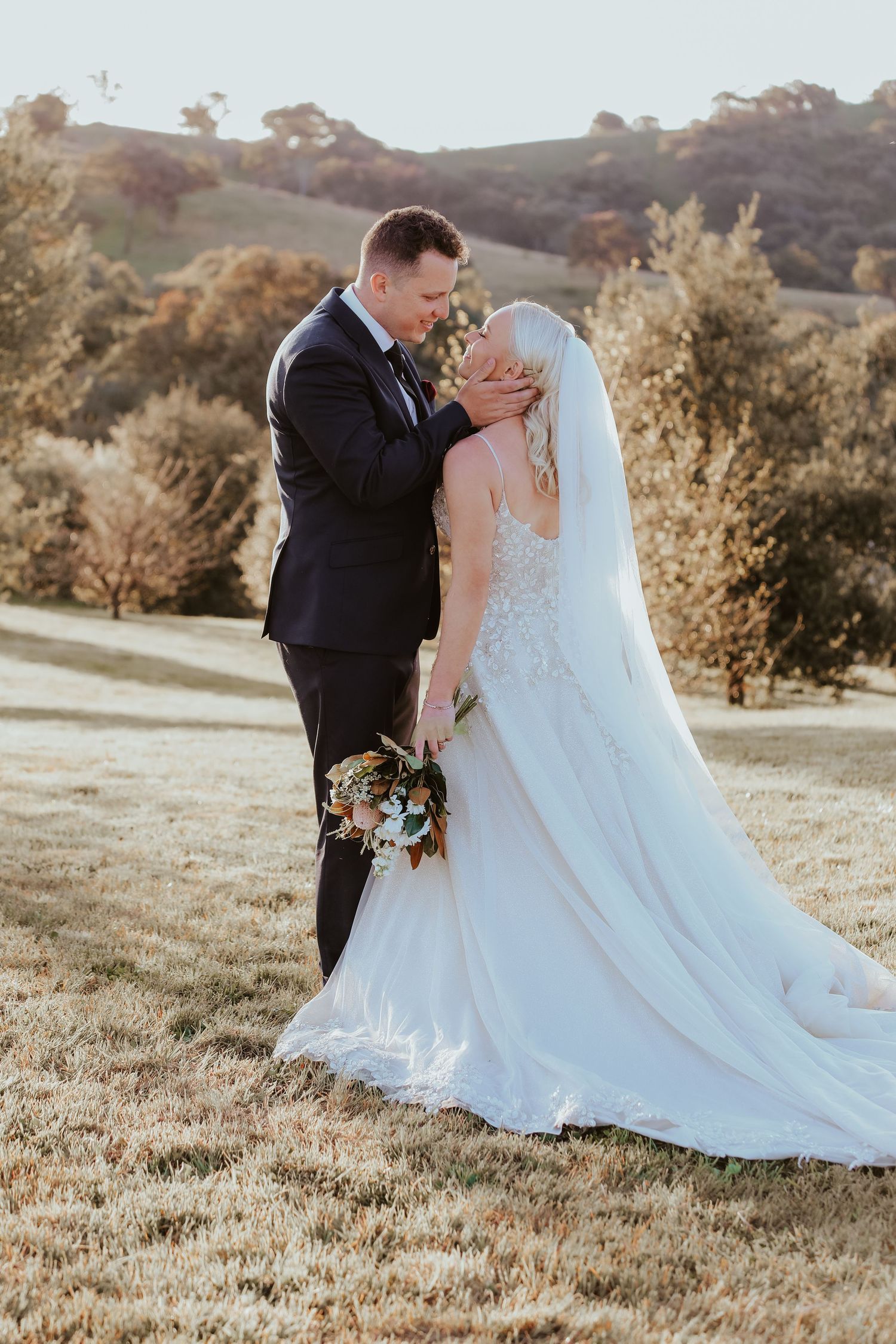 Wedding couple poses in scenic field with white dress trailing behind.
