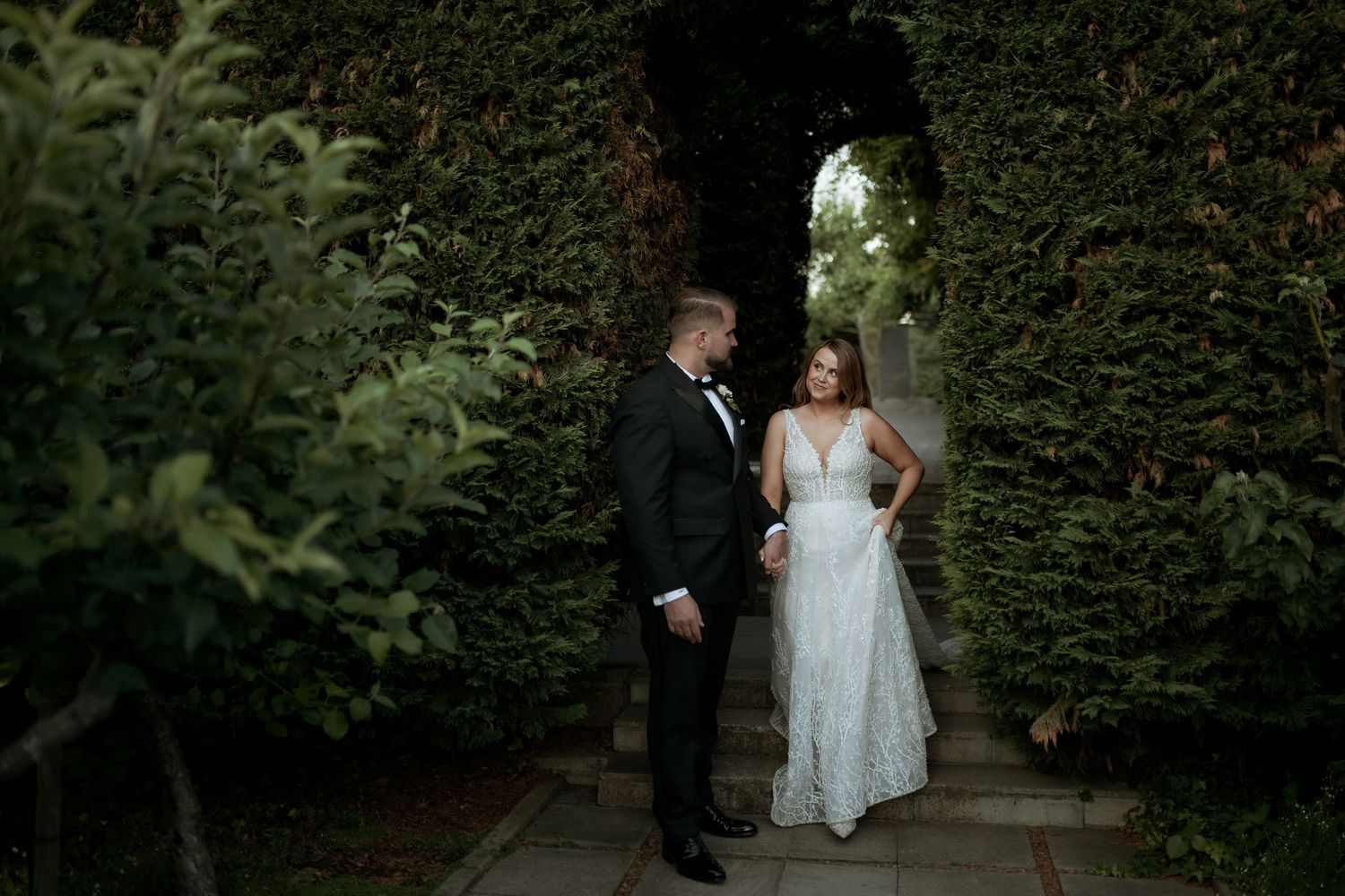 Bride in lace gown and groom in tuxedo stand together in a romantic garden archway of lush greenery.