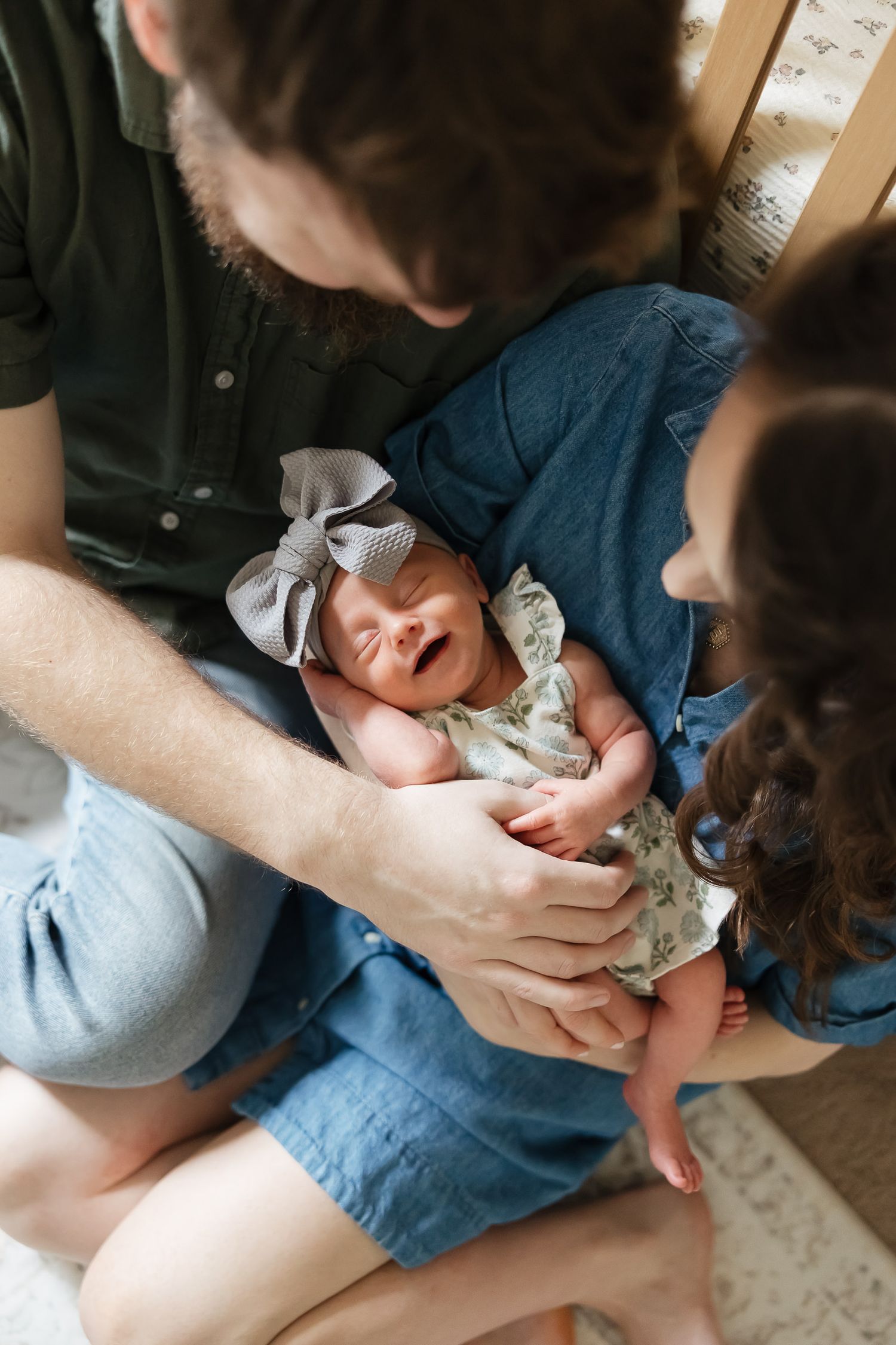 new parents sitting on floor infront of crib holding baby girl smirking up at camera as she sleeps overland park photographer
