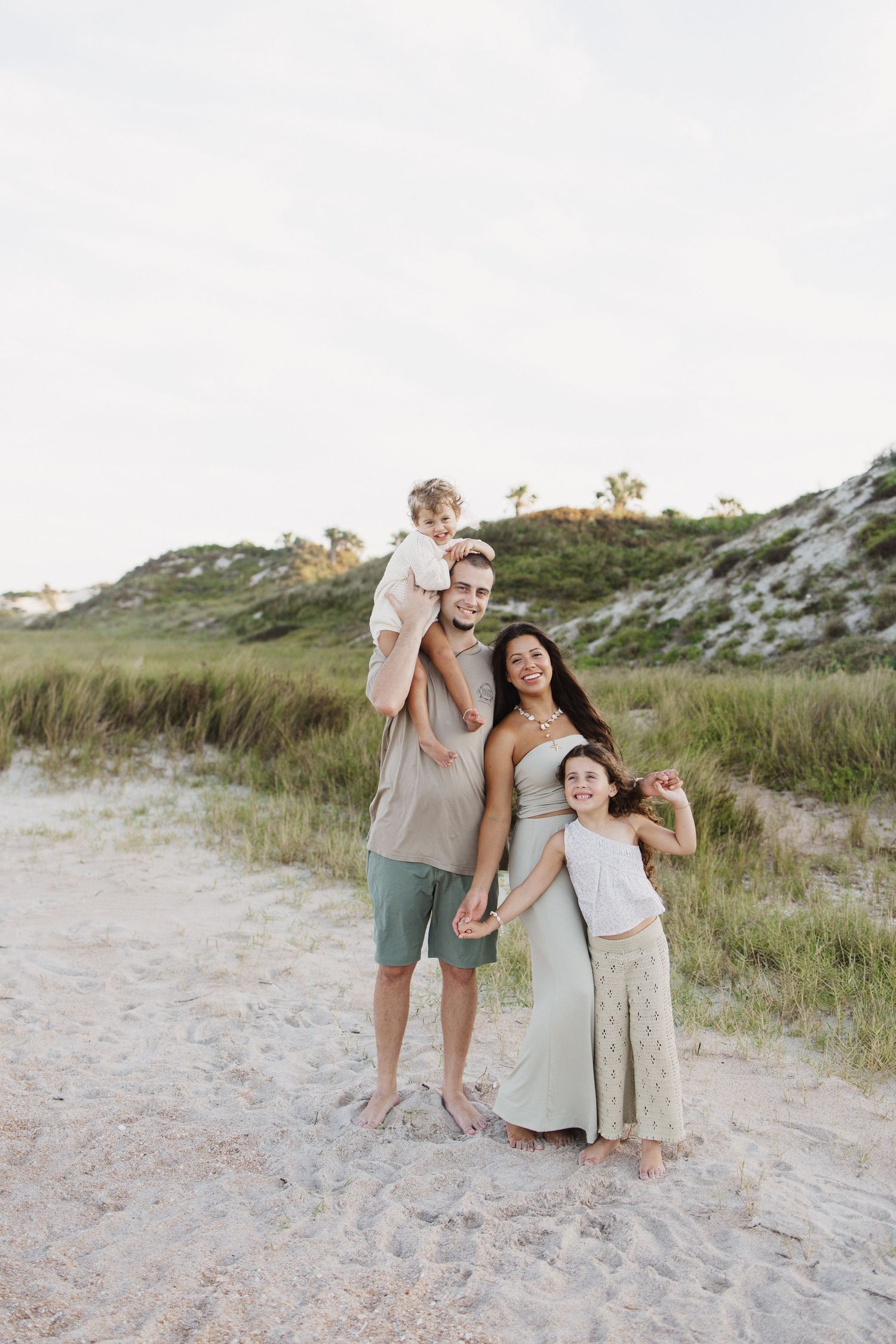 Happy family of four posing on sandy beach with green dunes and cloudy sky in background.
