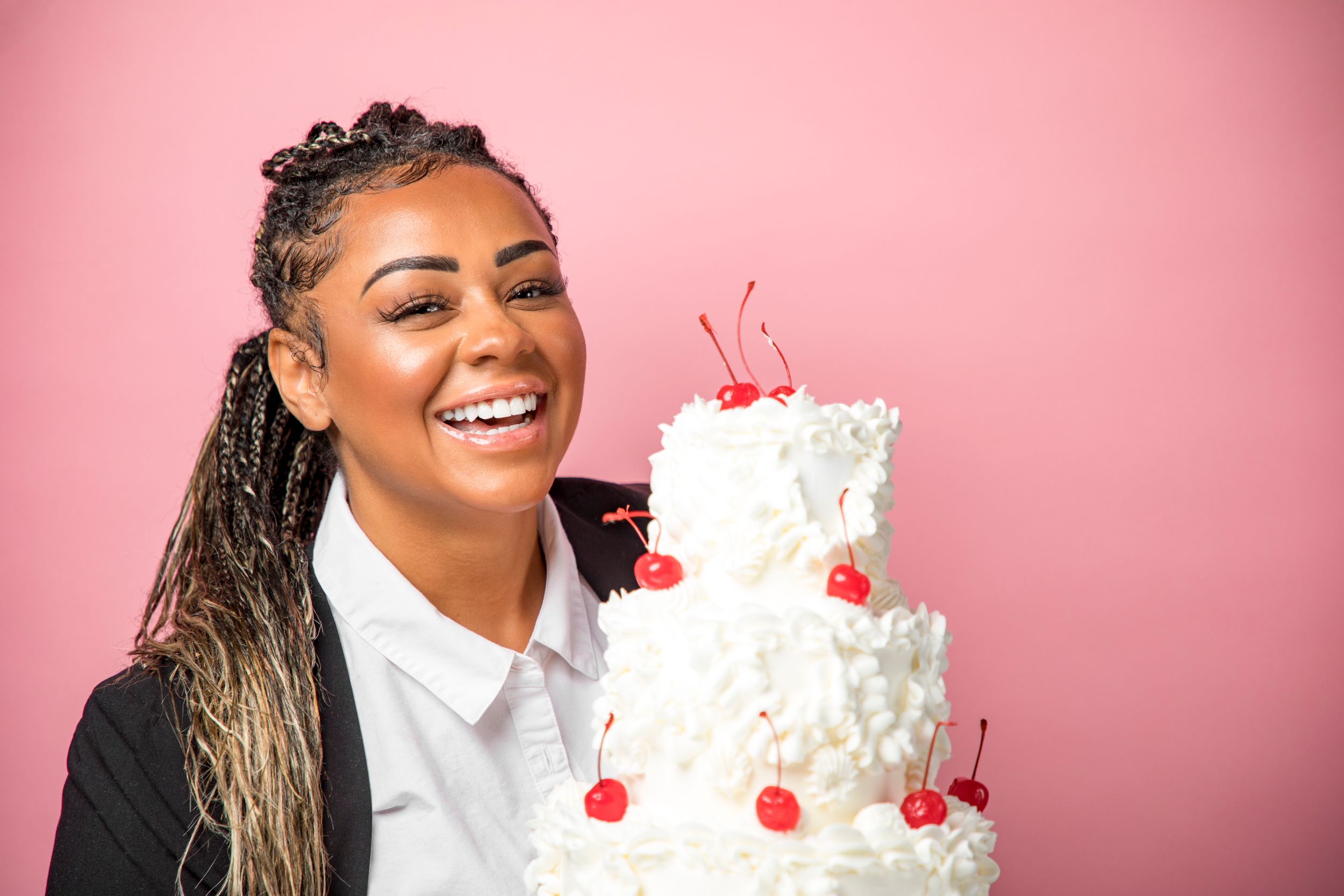 Woman in white shirt smiling next to elegant white wedding cake with red cherries.
