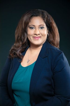 Professional headshot of someone wearing a navy blazer and teal blouse against dark background.