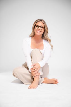 Person in white cardigan and beige pants sitting cross-legged on white background.