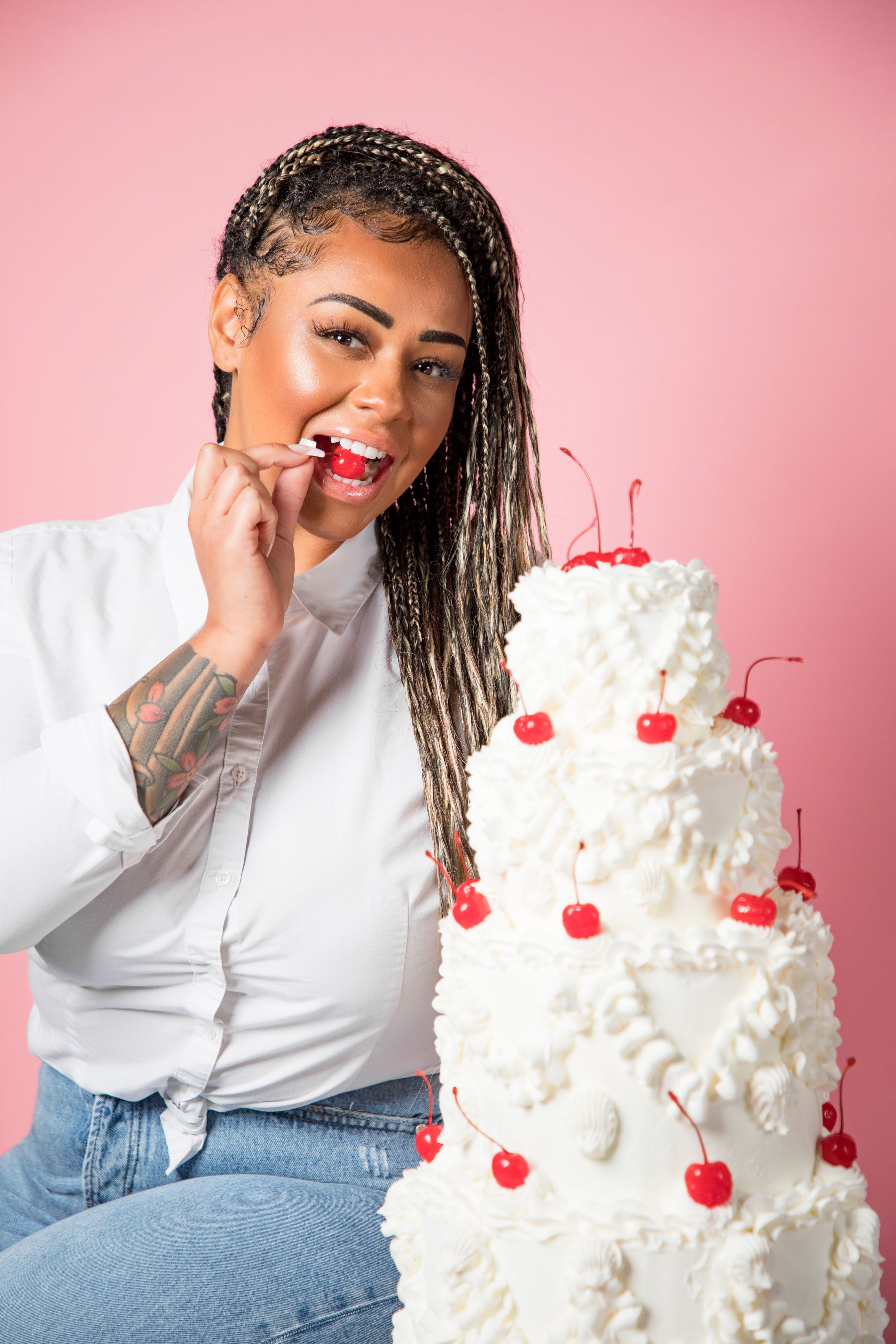 Woman posing playfully with three-tiered white wedding cake against pink backdrop.