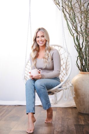 Person sitting in macrame hanging chair holding white coffee mug.