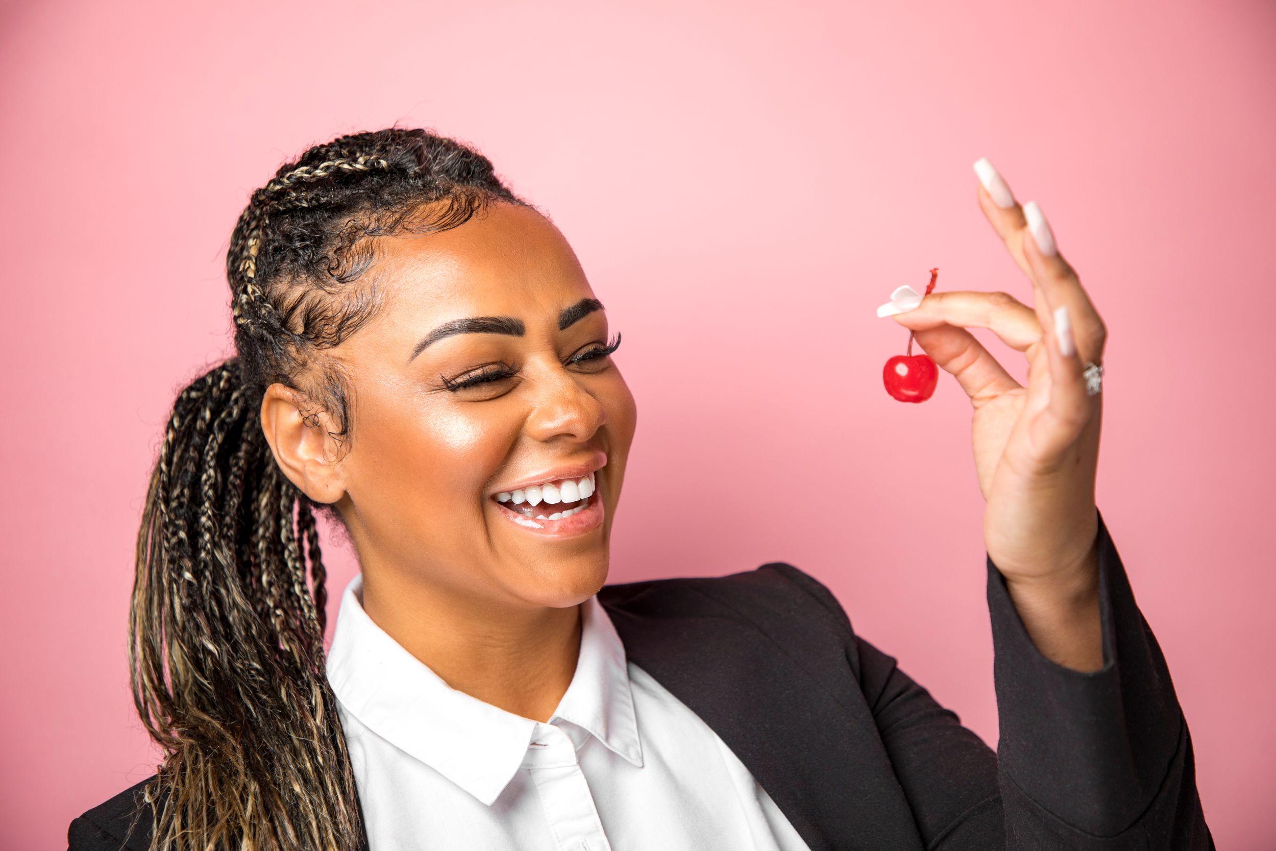 Woman laughing while holding cherry from wedding cake decoration against pink background.