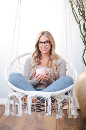 Person wearing glasses and gray sweater relaxing in white macrame hanging chair.