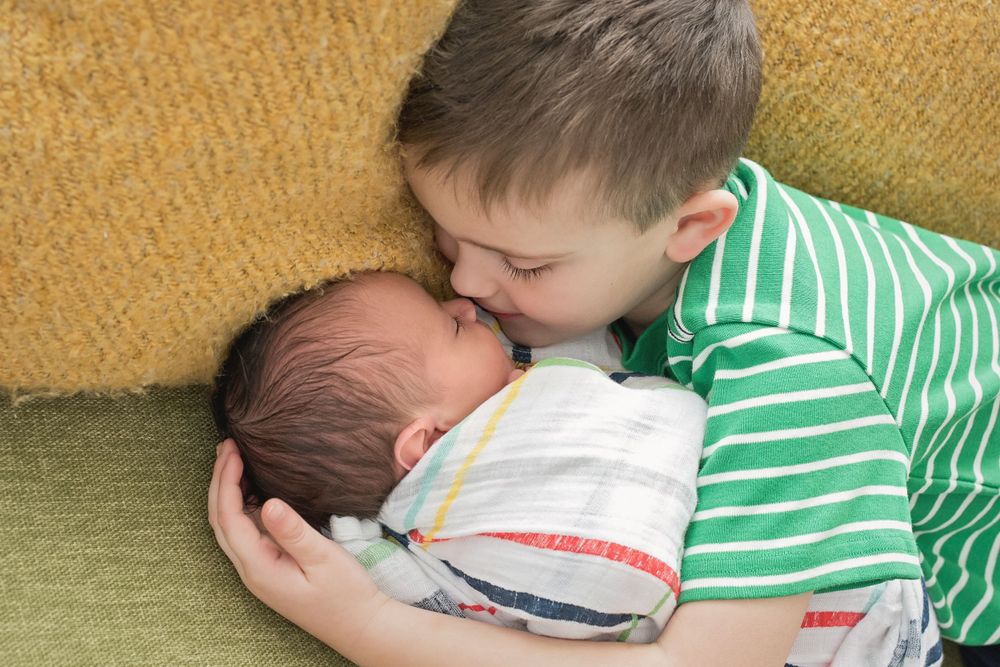 Young child gently kisses newborn sibling on yellow couch.