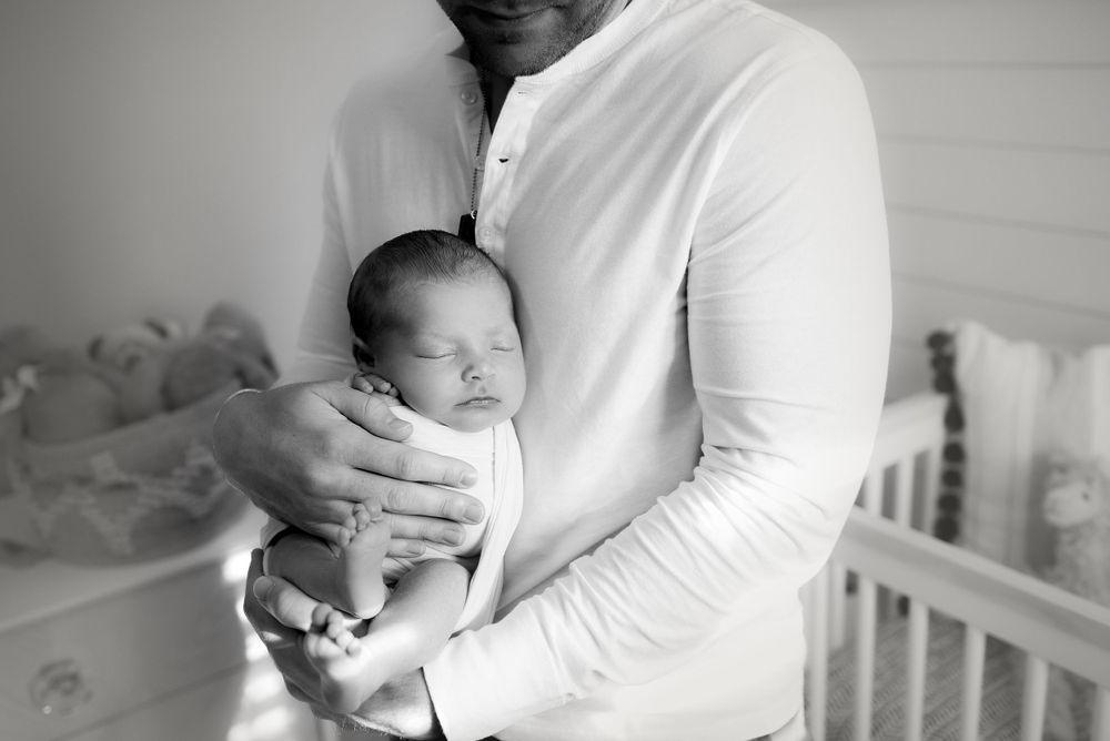 A tender black and white photo of an infant being cradled in someone's arms near a crib.