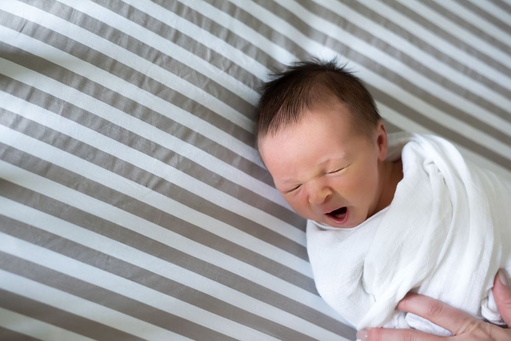 Newborn baby in white sleeper yawning on striped bedding.