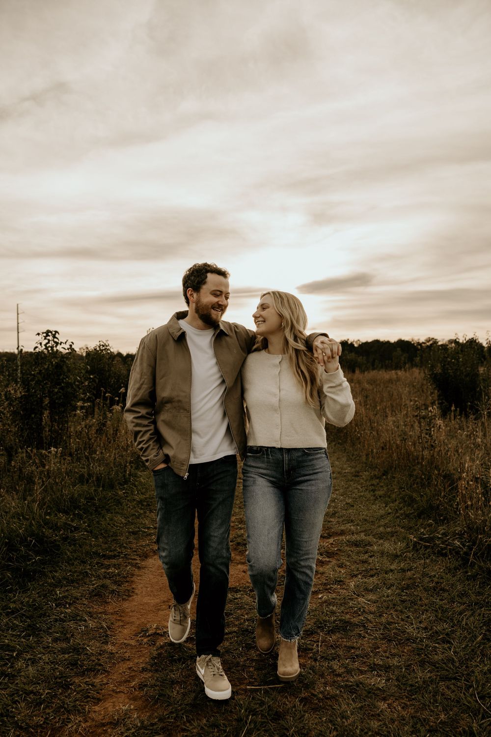 A couple walks together smiling on a dirt path through a field during golden hour.