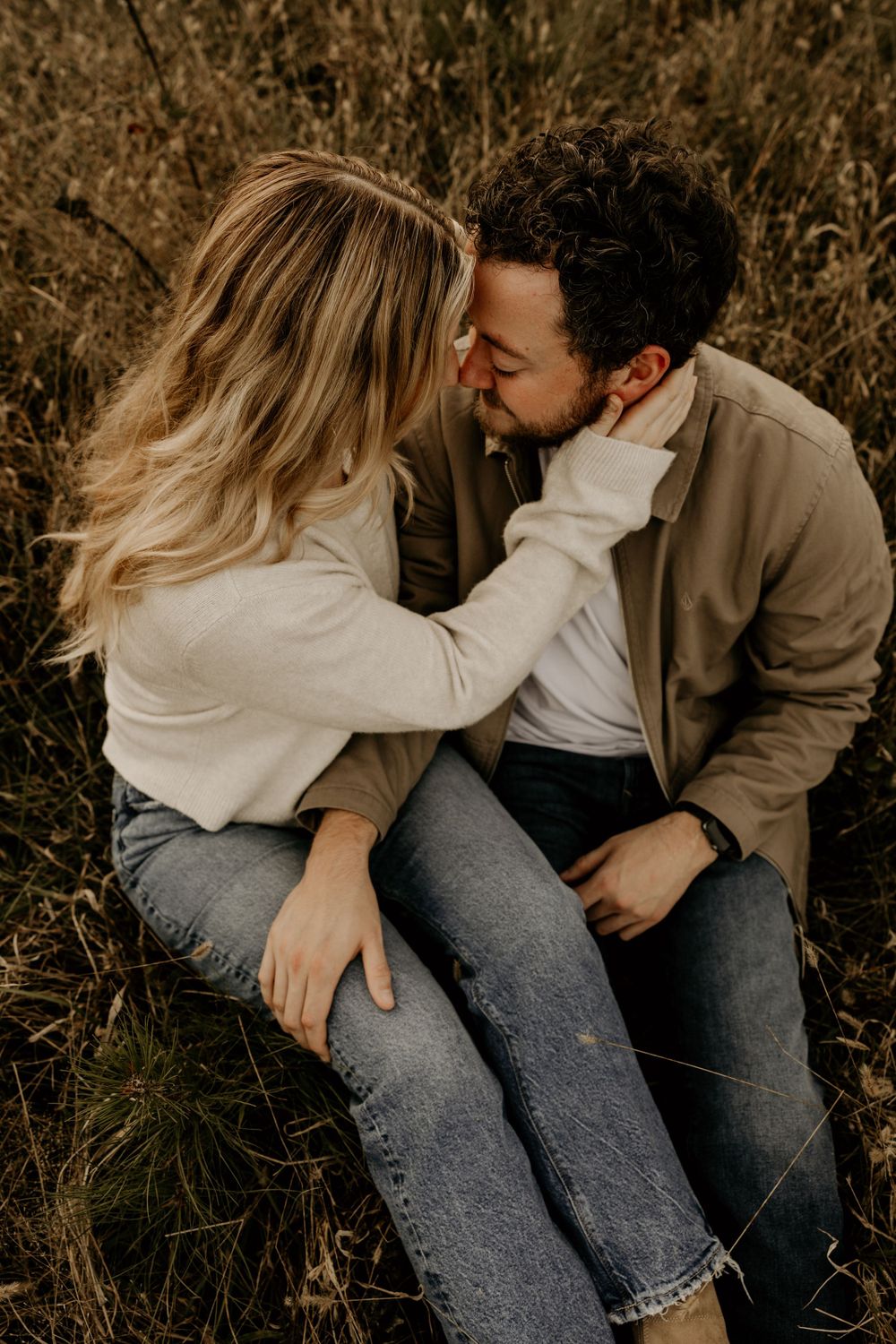 Couple in cozy sweaters embracing while sitting in tall autumn grass during golden hour.