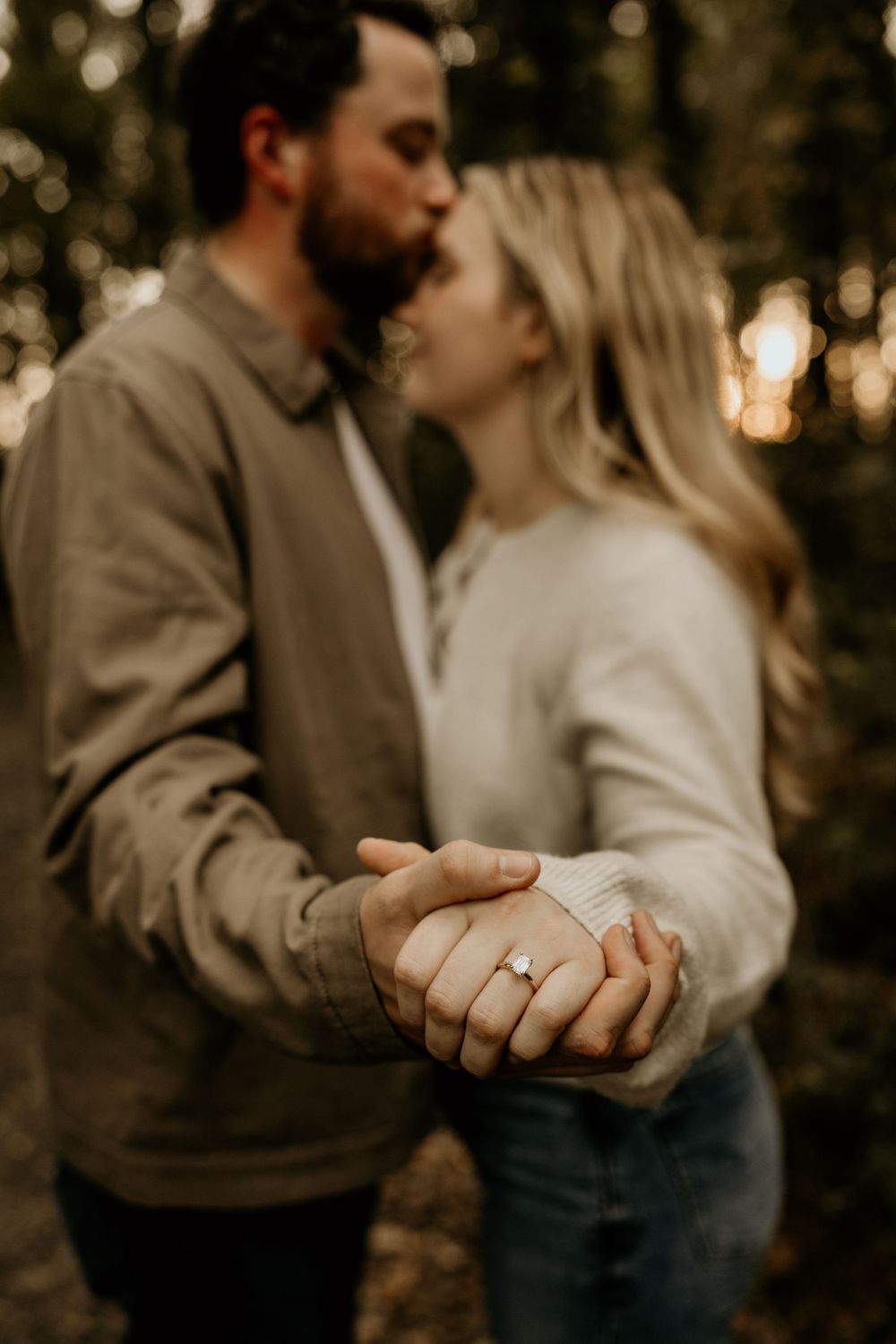 An engaged couple holding hands with focus on the ring during a romantic forest photoshoot at sunset.